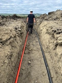 A person standing in a narrow trench with black and red cables running along the ground.