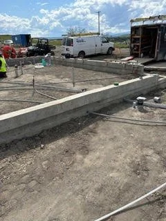 Construction site with concrete foundation being laid, construction workers, and vehicles including a white van, under a partly cloudy sky.
