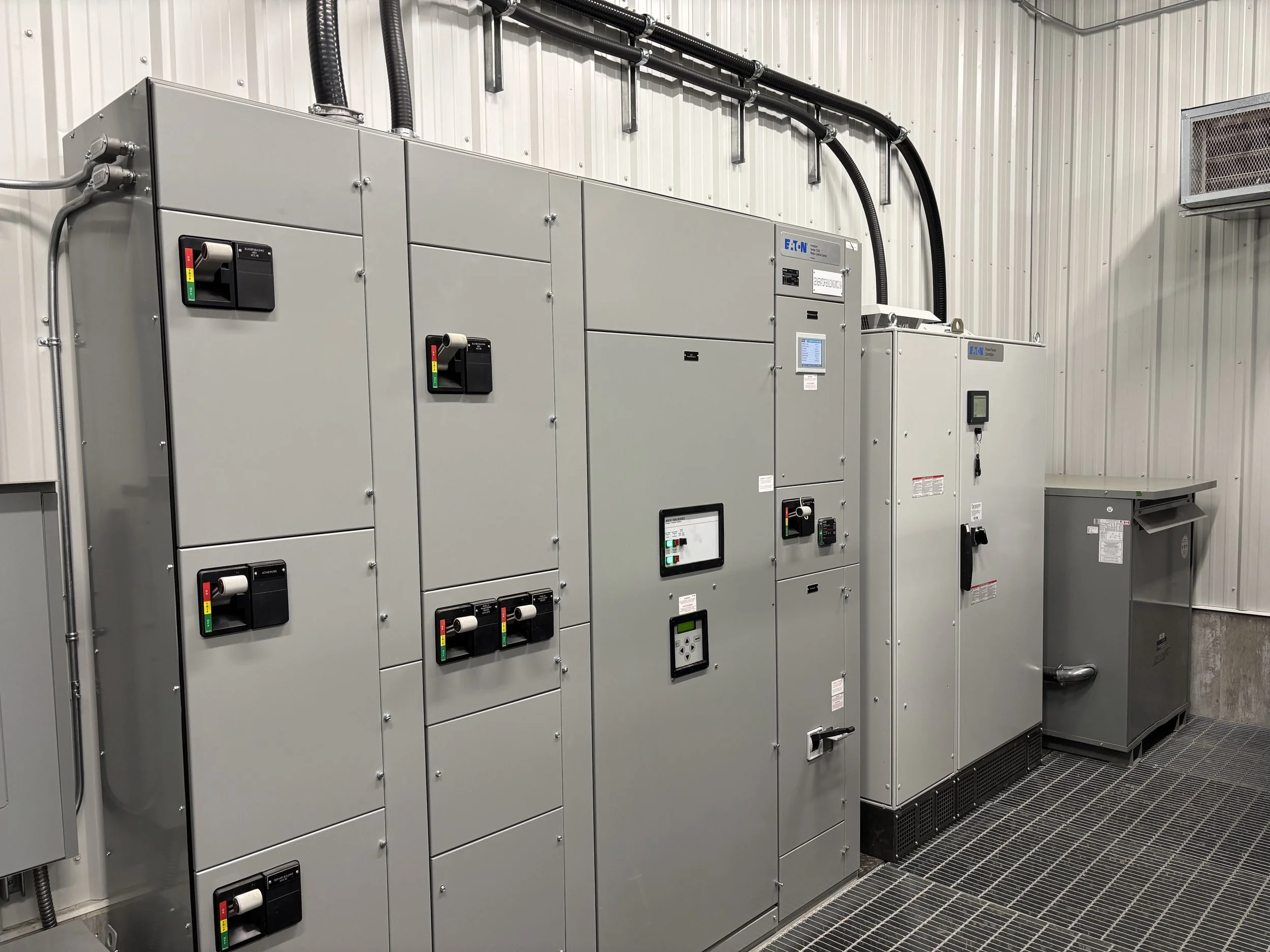 Industrial electrical equipment, including control panels and circuit breaker switches, installed on a metal wall in a facility, with wiring and ventilation.