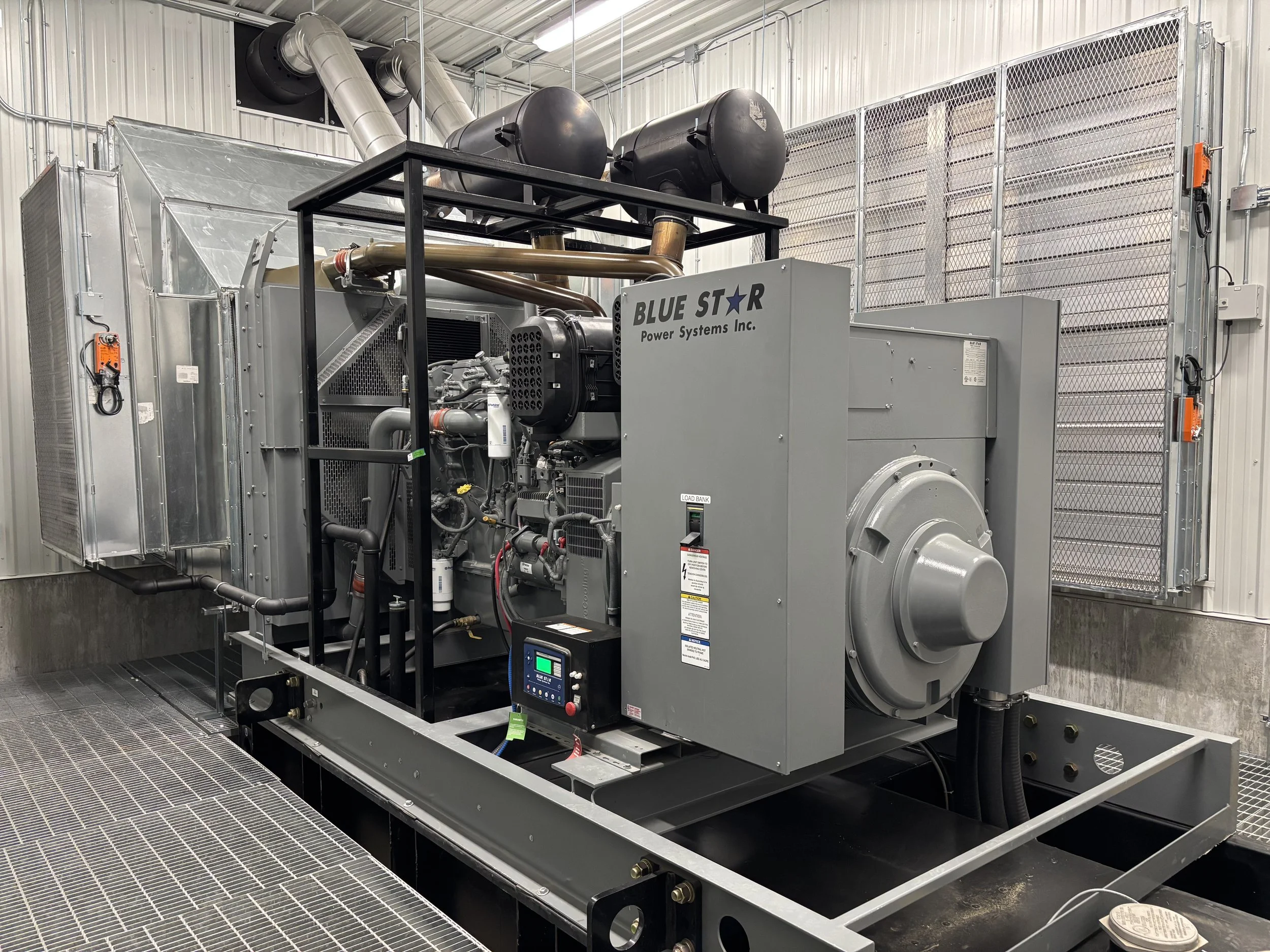 Large industrial generator labeled 'Blue Star Power Systems Inc.' situated in a clean, industrial room with metallic walls and grated flooring.