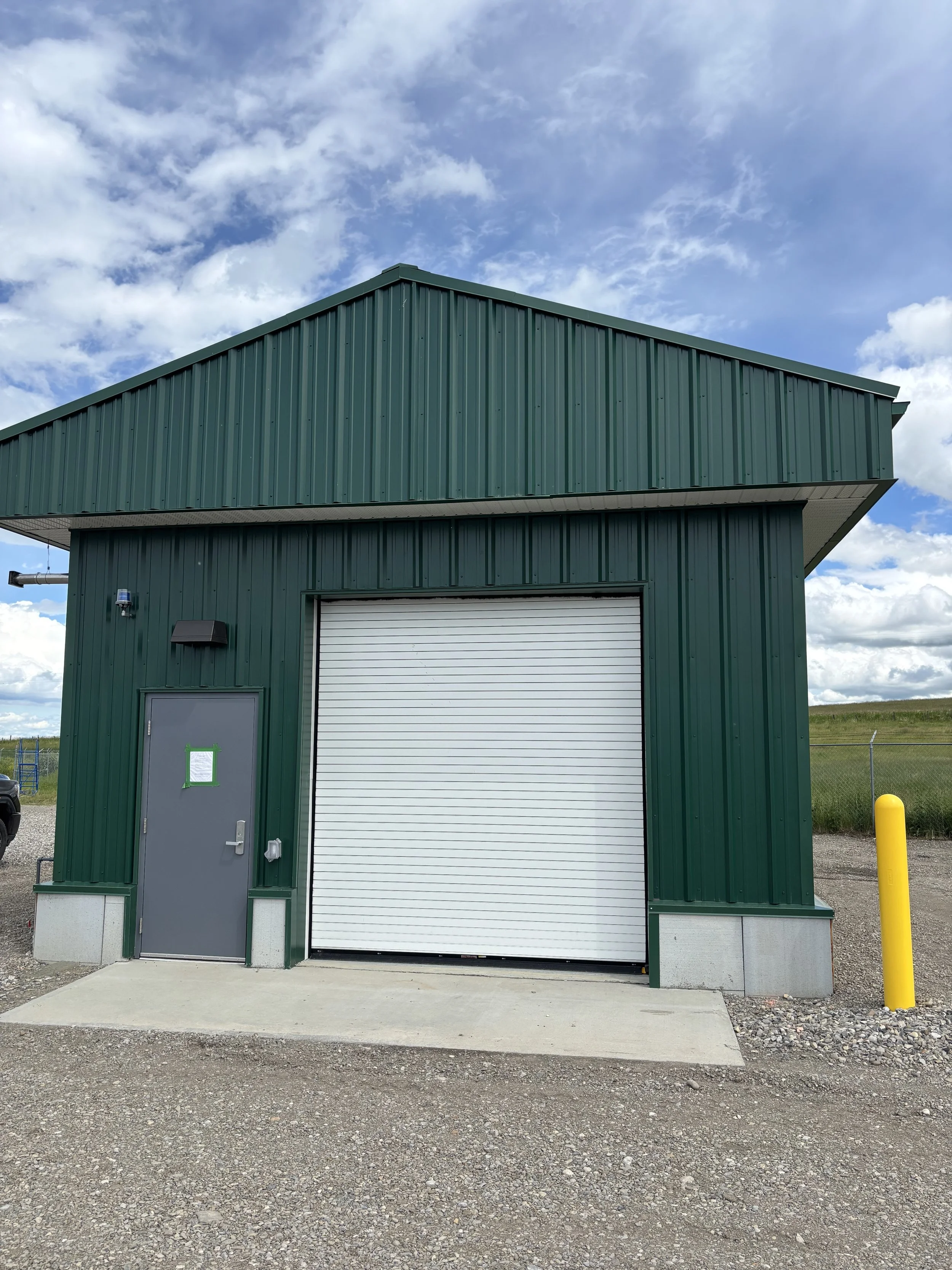 A green industrial building with a metal exterior and a white roll-up garage door, situated on a gravel lot with a concrete pad in front, under a partly cloudy sky.