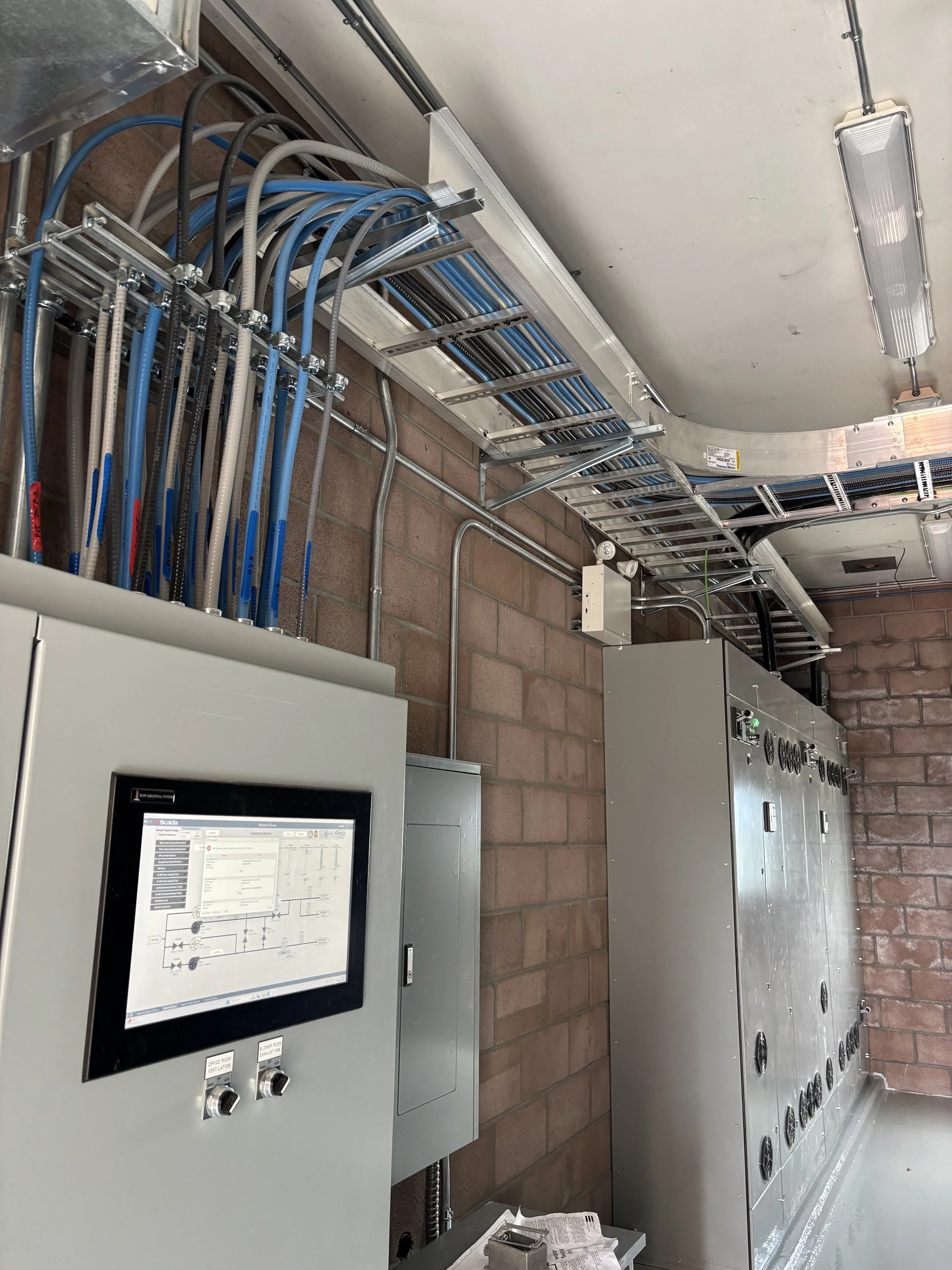 A row of gray electrical control panels with a touchscreen interface on the front, mounted on a wall with exposed brick. Extensive conduit wiring runs along the ceiling and wall, connected to various components. There are industrial ceiling lights an