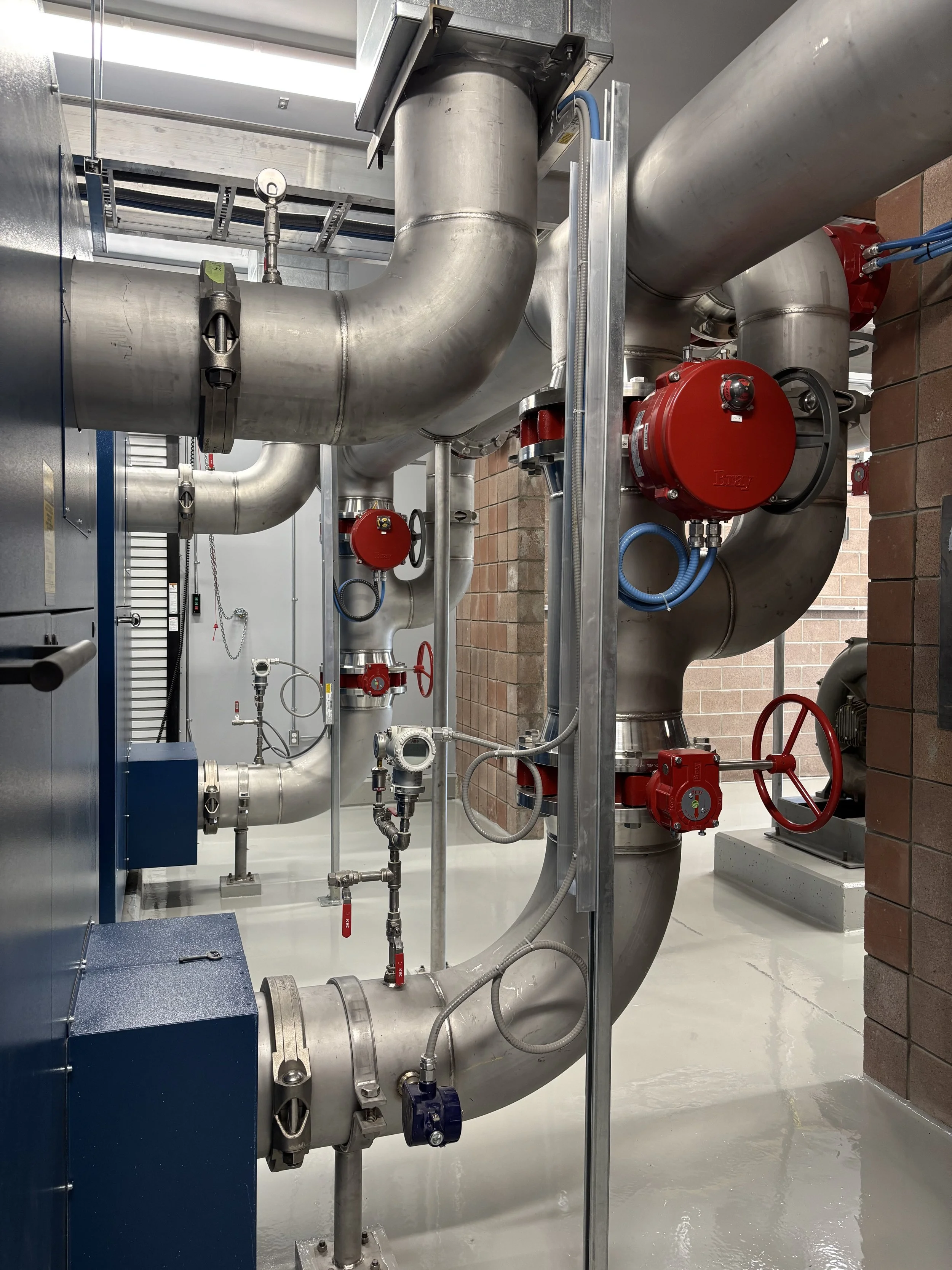 Industrial piping system with large metal pipes, valves, and red components inside a building with brick wall and concrete floor.