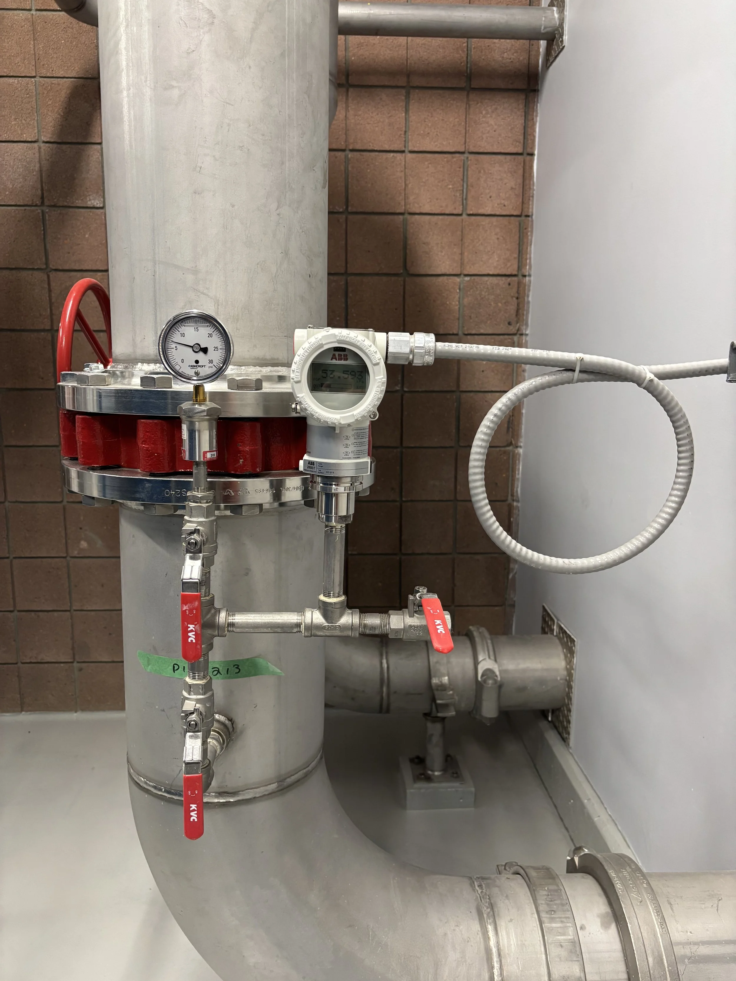 An industrial piping system with valves, a pressure gauge, and a digital display, installed against a brick wall.