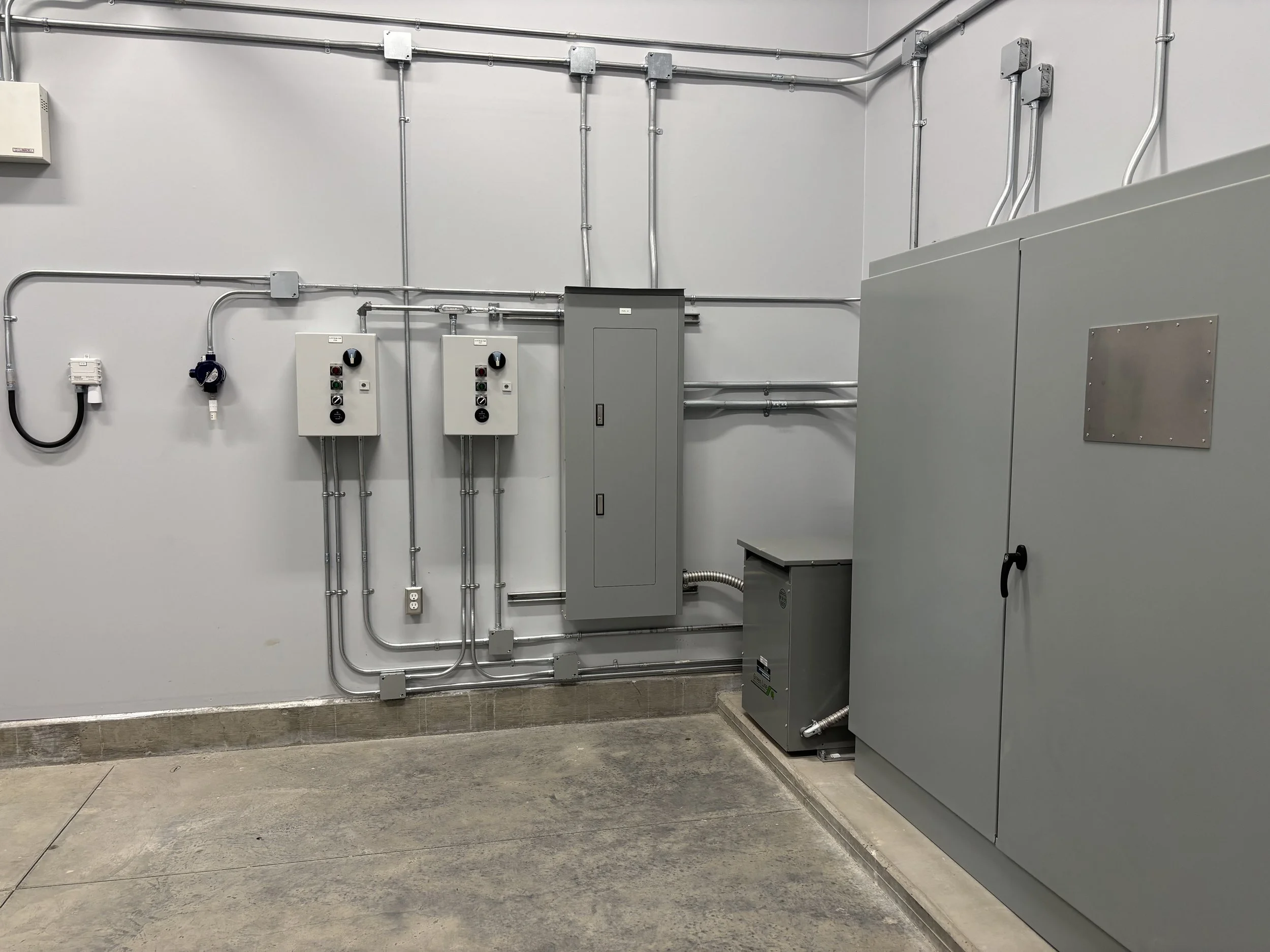 Electrical wiring and control panels mounted on a plain white wall in an industrial or commercial setting.