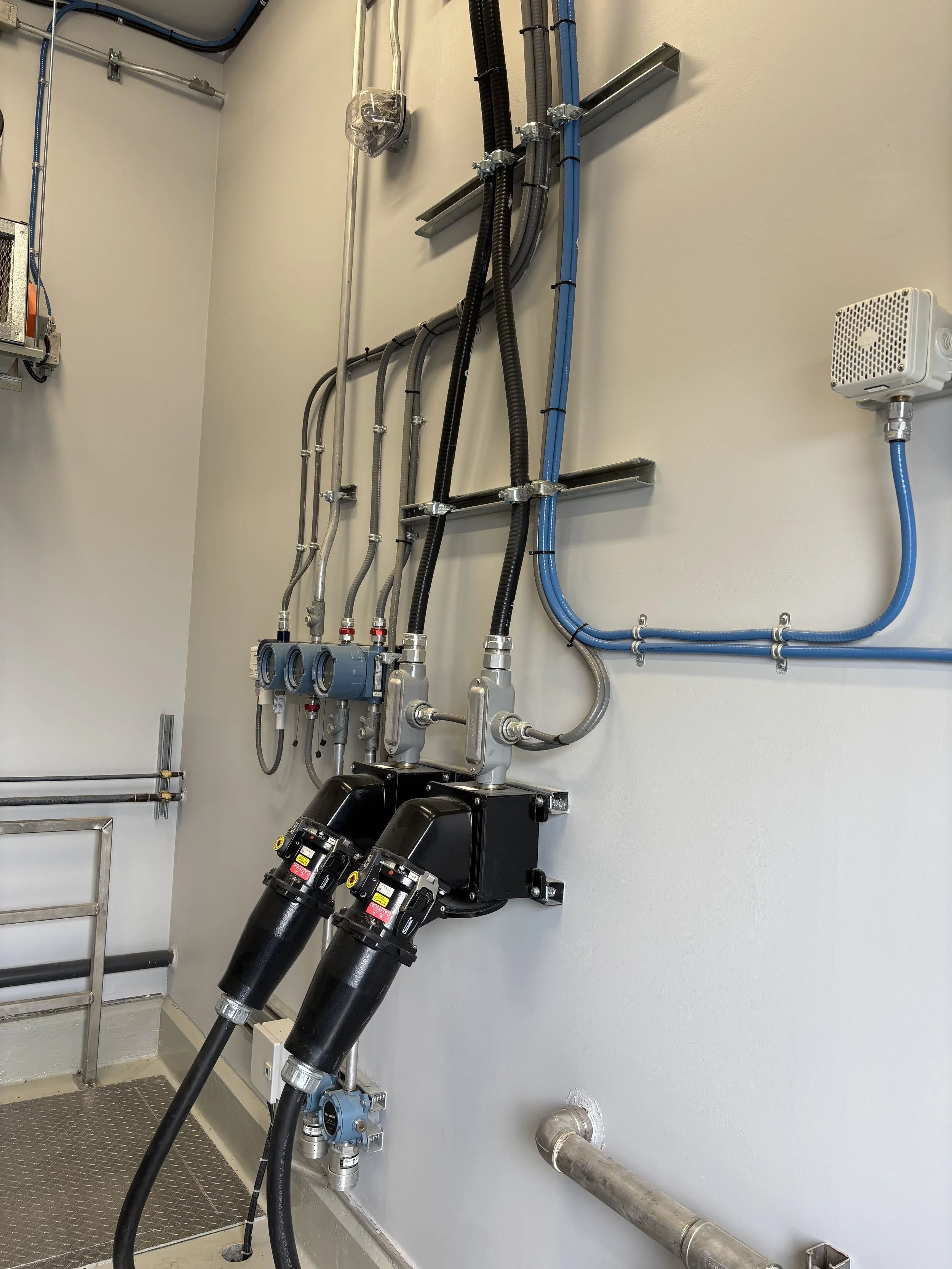Industrial piping and meters with wiring and electrical components attached to a light-colored wall in a utility or mechanical room.