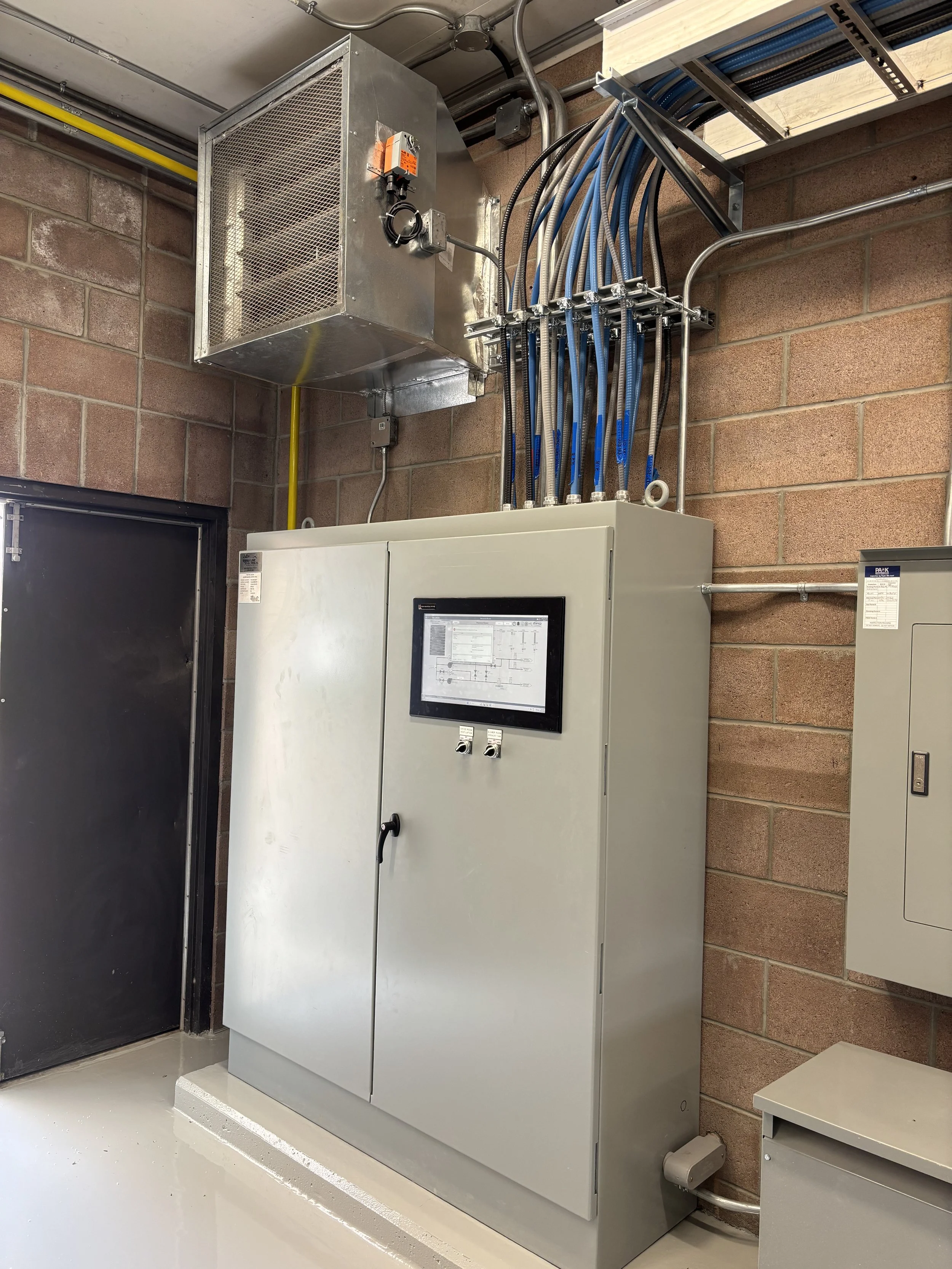 Electrical control panel with wiring and components mounted on a brick wall in an industrial setting.