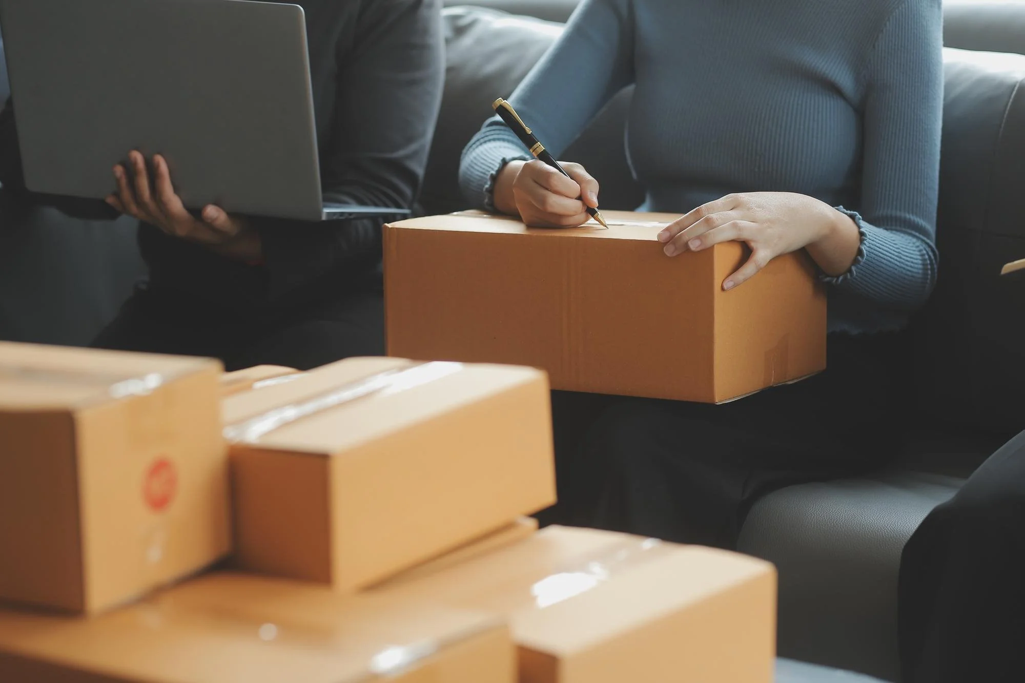 People sitting on a couch, handling packages and writing on a box, with more boxes in the foreground.