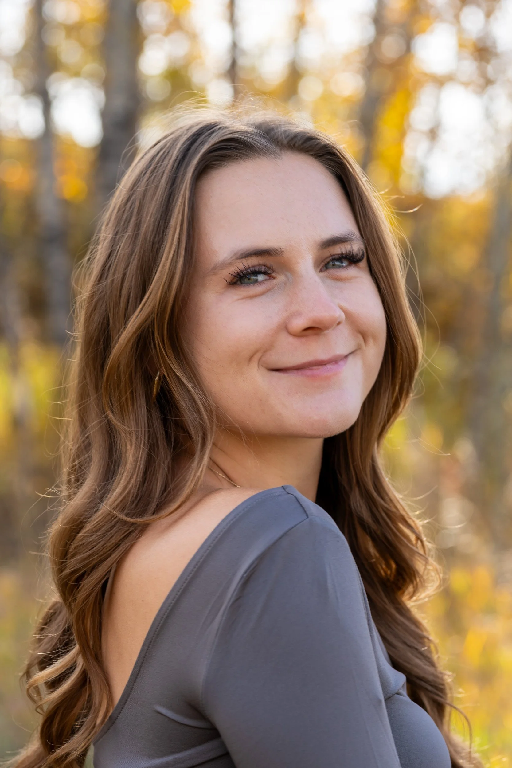 A woman with long, wavy brown hair and blue eyes smiling outdoors in front of a background of autumn leaves and trees.