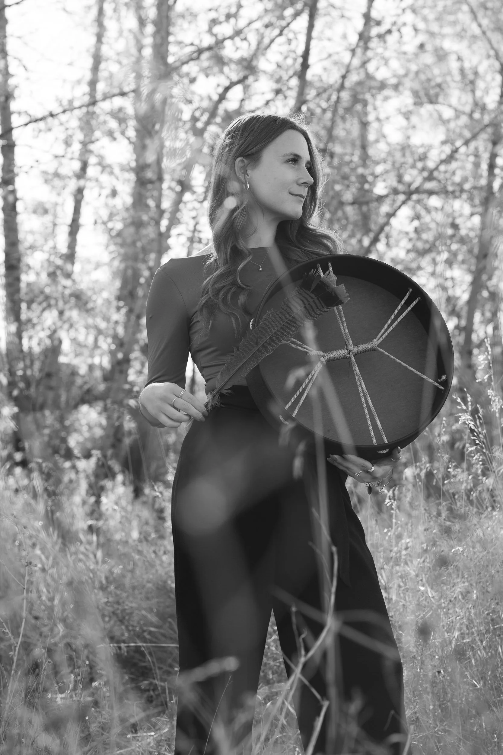 A woman in a dress holding a healing drum, standing outdoors in a wooded area.