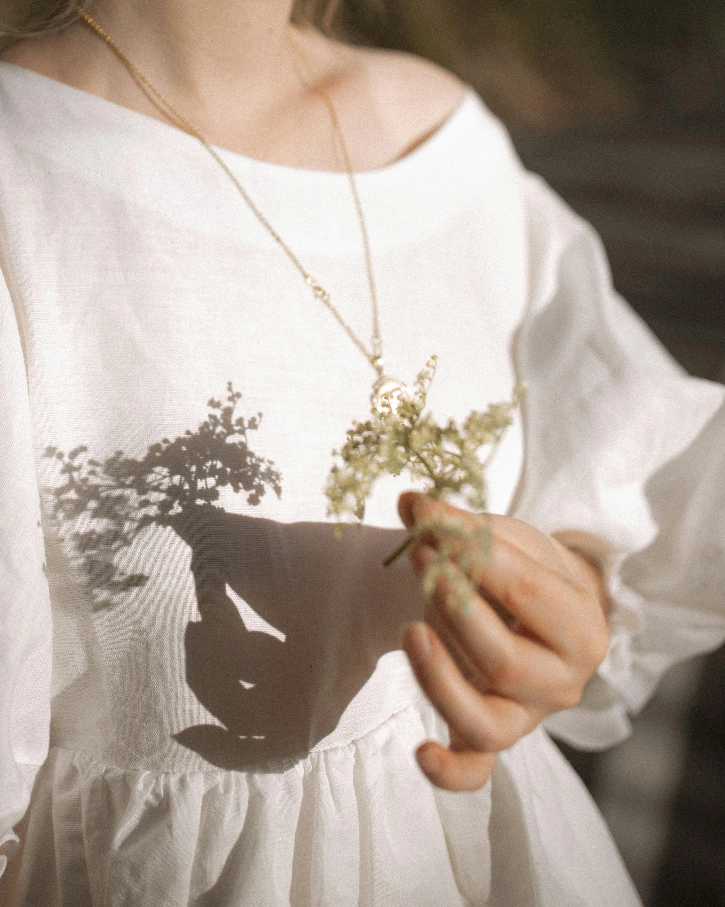 Close-up of a woman wearing a white dress and delicate gold necklace, holding a small bouquet, with a shadow of her plant cast on her dress.