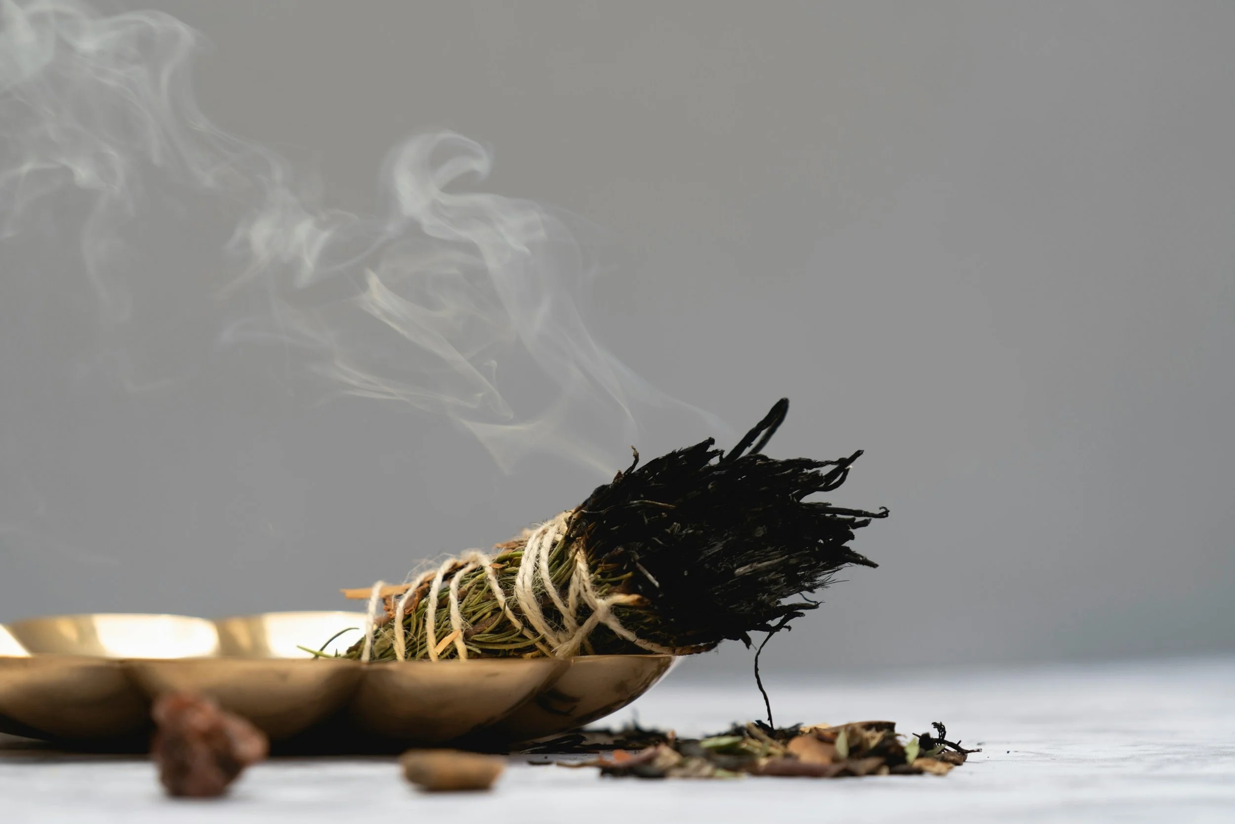 A bundle of burning sage tied with twine, releasing smoke, placed on a metallic bowl.