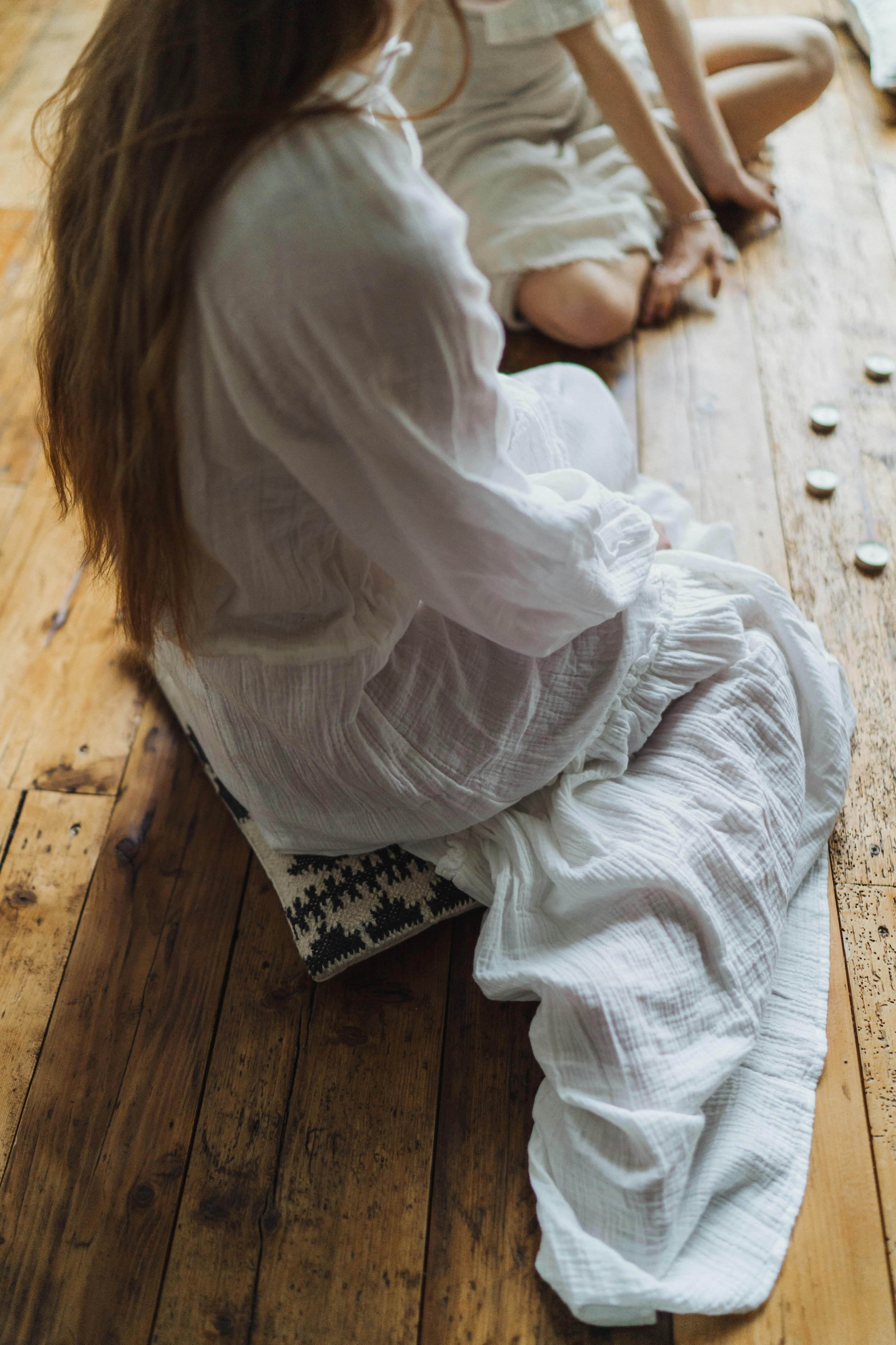 A person dressed in white, likely a woman, sitting on a wooden floor with their legs folded and hands resting on their lap. The image captures their side profile with long hair and a henna tattoo on the hand, detailed with white candles placed in a line on the floor nearby.