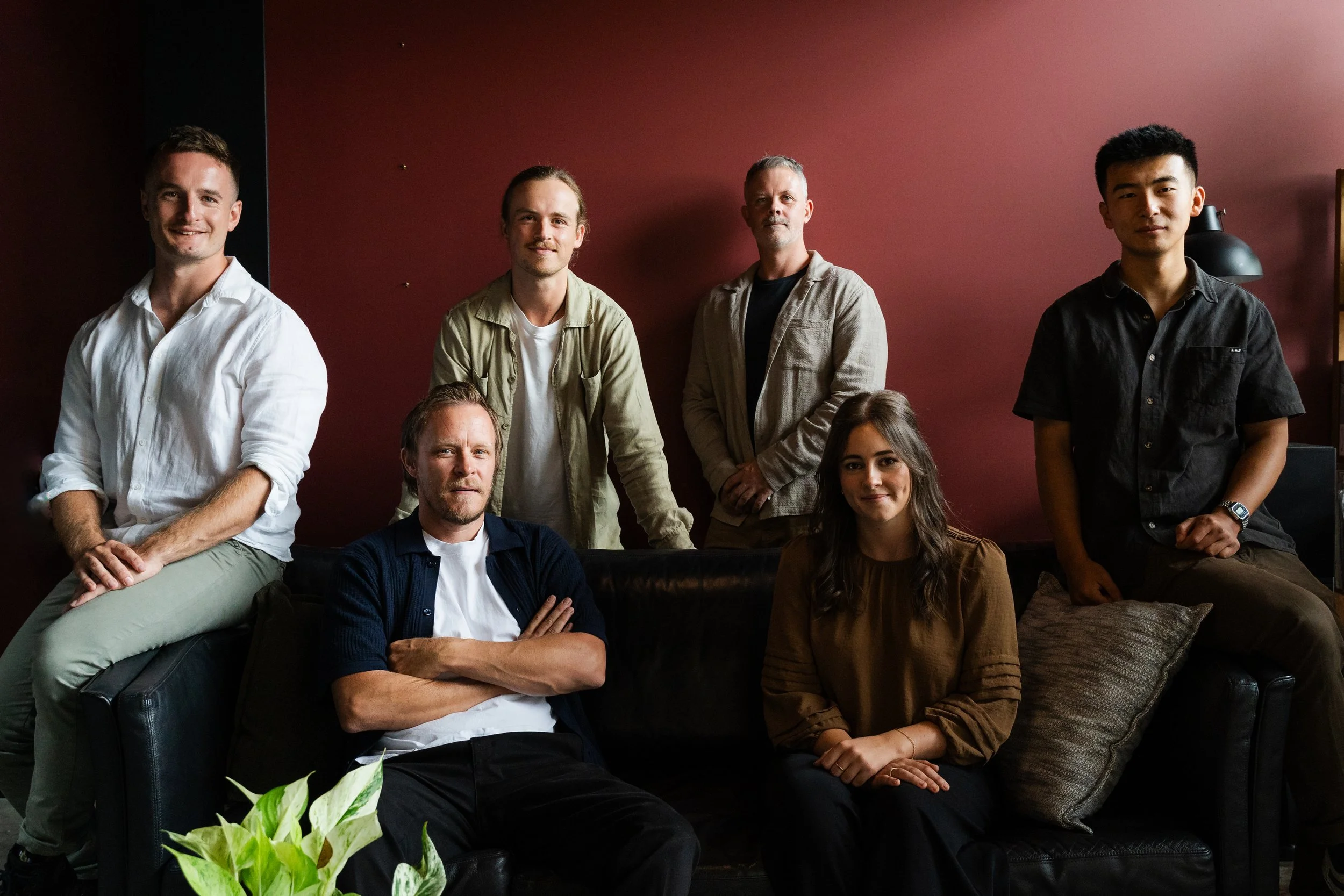 A group of seven diverse young adults, four men and three women, gathered in a cozy indoor setting with a dark maroon wall and warm lighting.