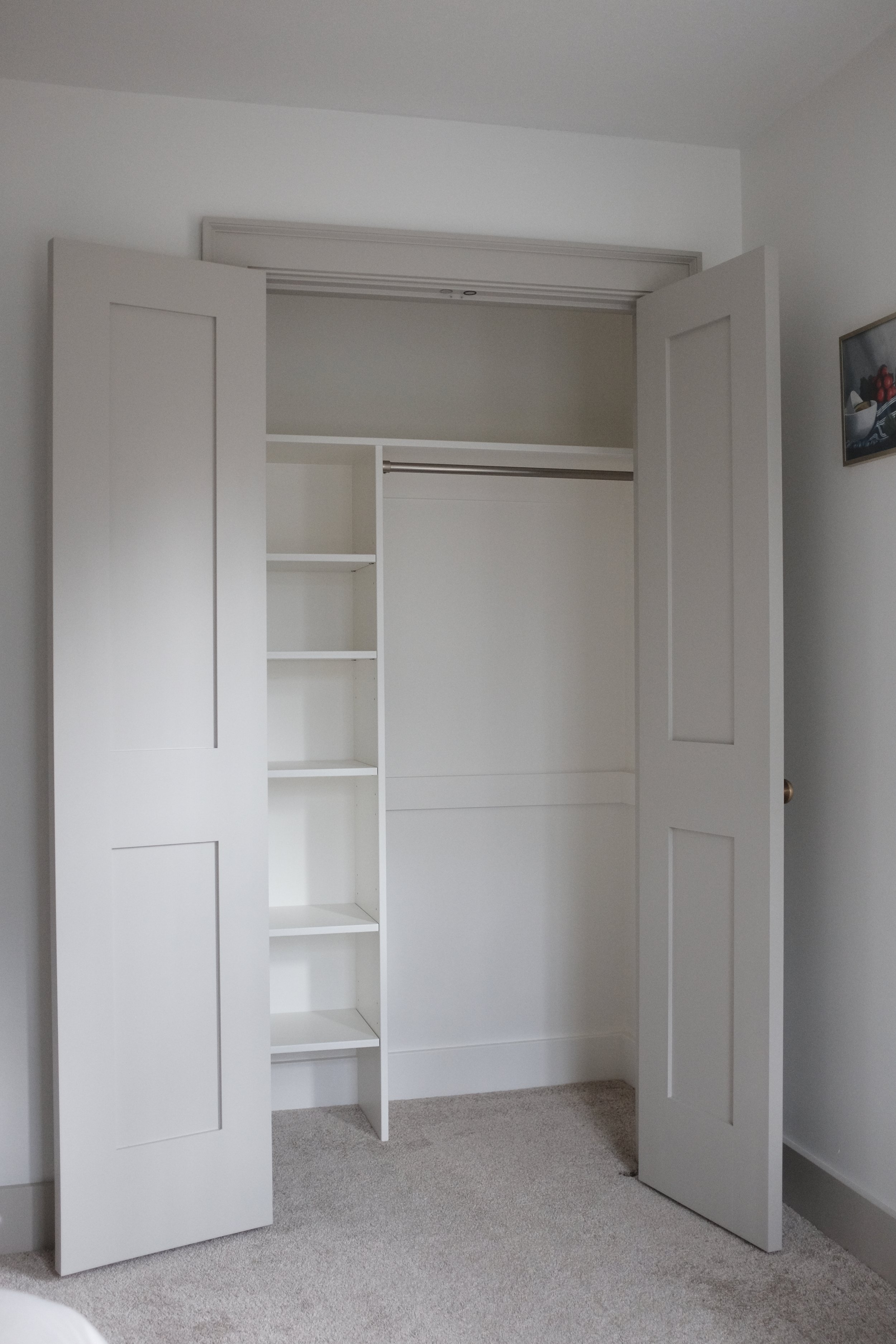 Open closet with white sliding doors, shelves, and a hanging rod above a carpeted floor in a minimalistic room.