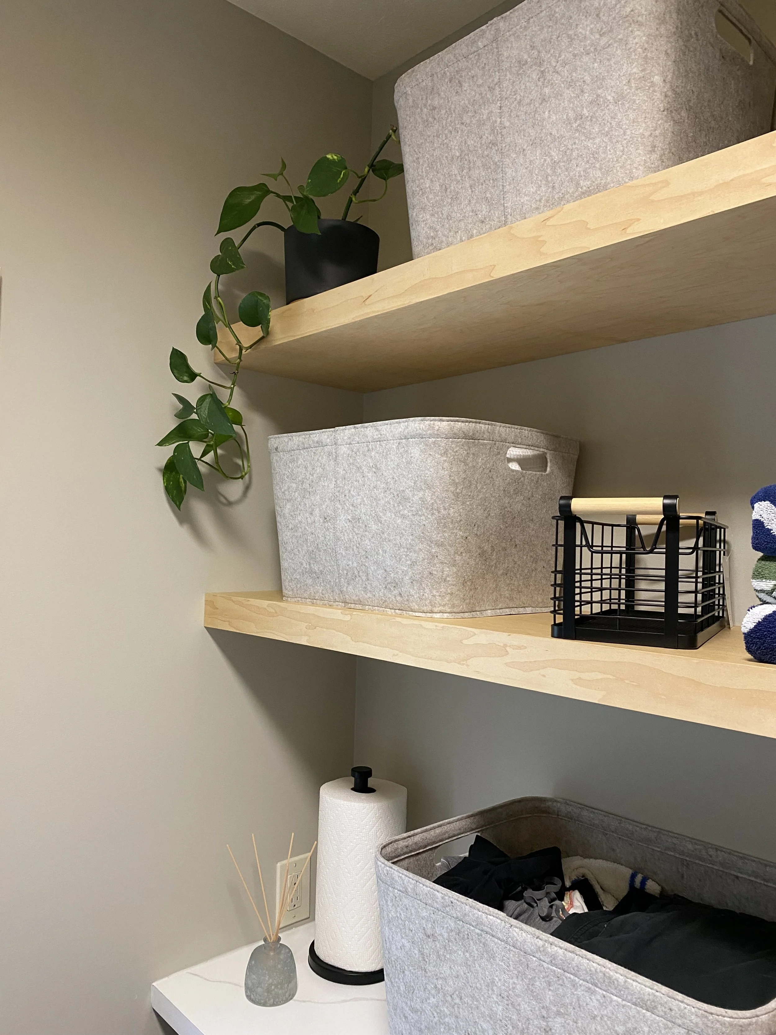Laundry room with wooden shelves, gray storage bins, paper towel holder, reed diffuser, black wire basket, and a potted plant.