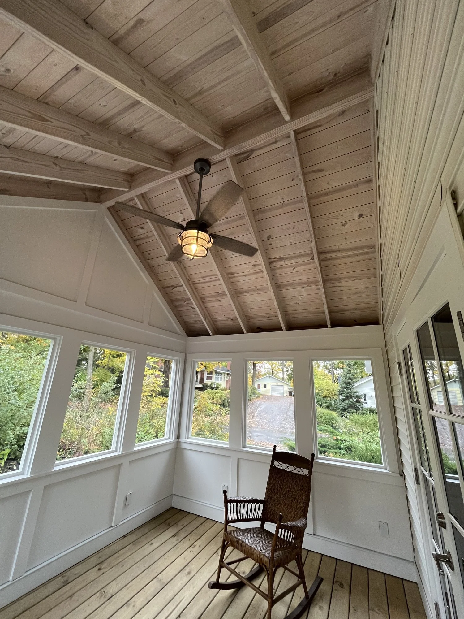 Enclosed porch with wooden ceiling, ceiling fan, large windows, and a wicker rocking chair.