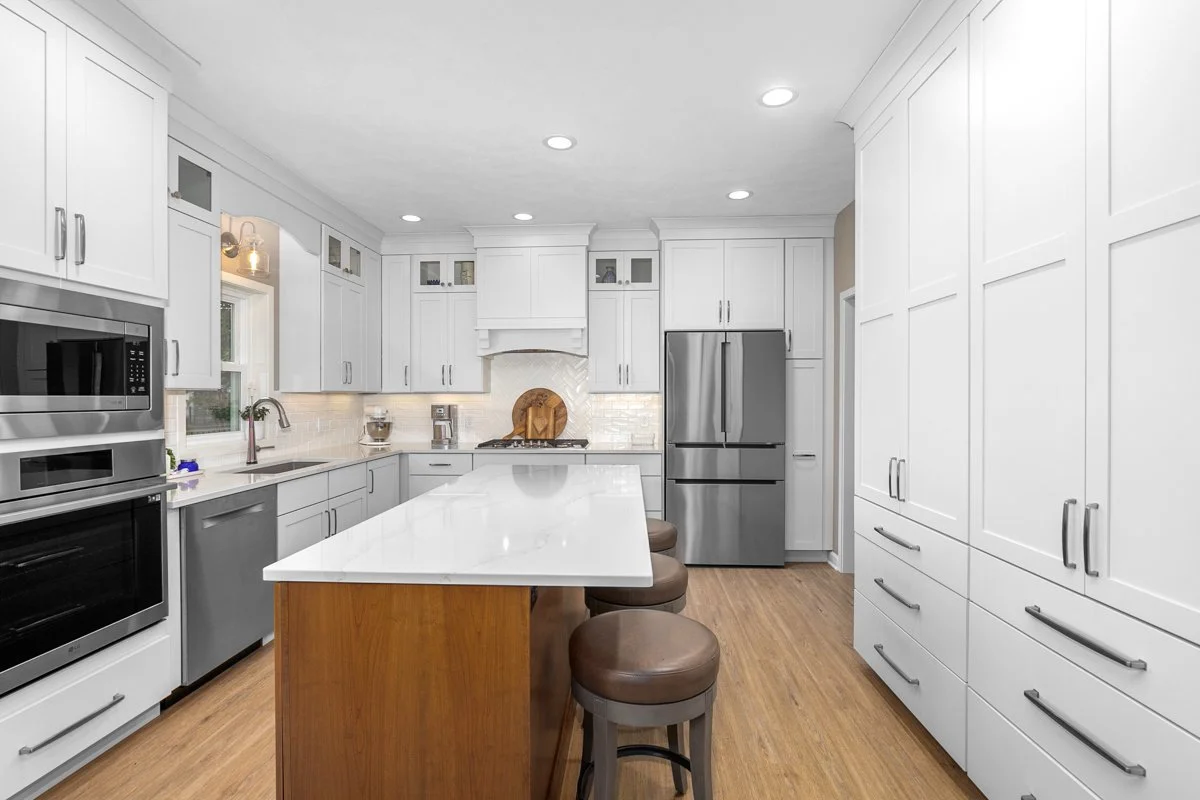 Modern white kitchen with stainless steel appliances, a large island, wooden stools, and wood flooring.