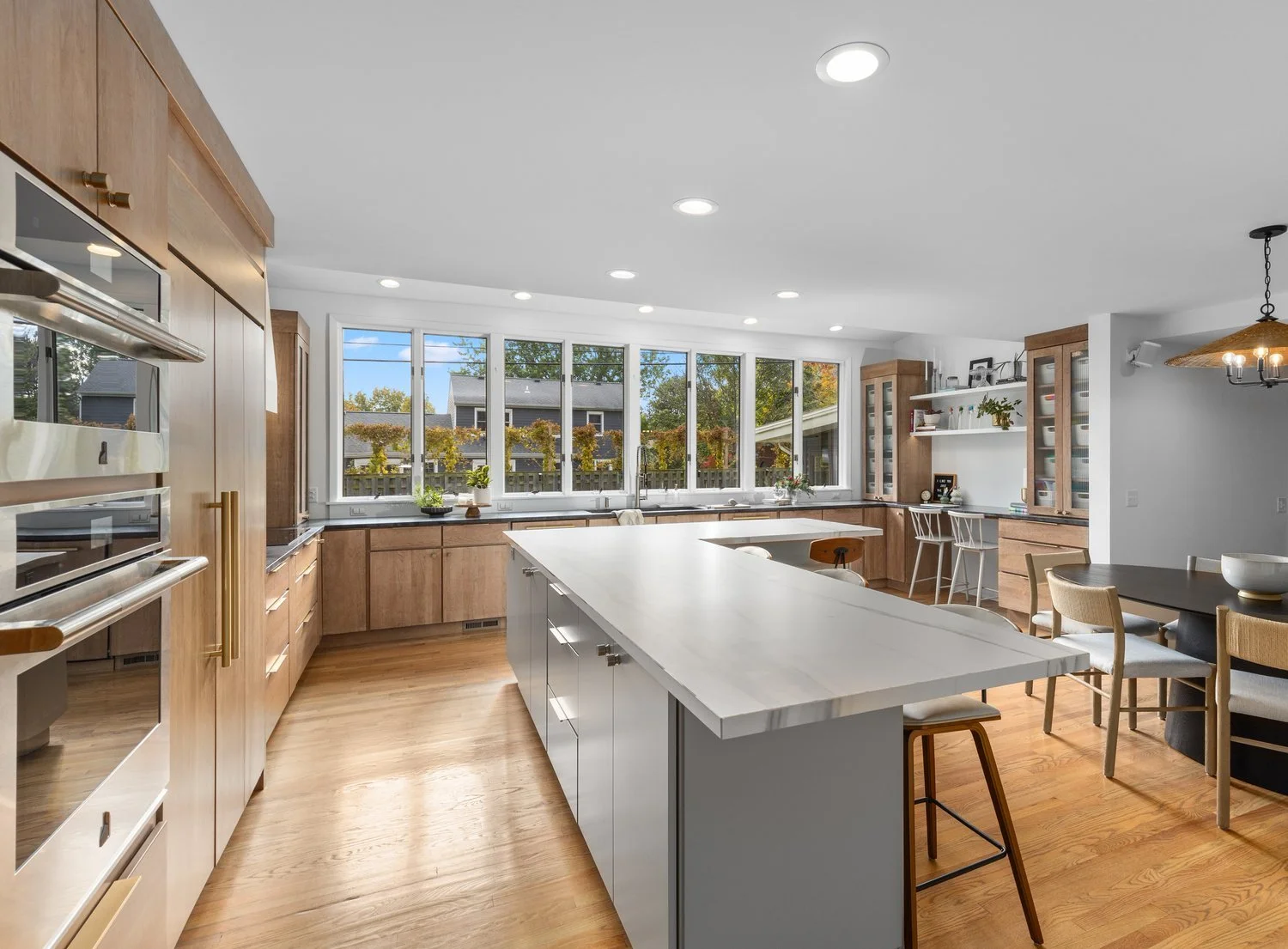 Modern kitchen with large island, wooden cabinets, and stainless steel appliances, featuring a dining area with chairs and scenic window views.