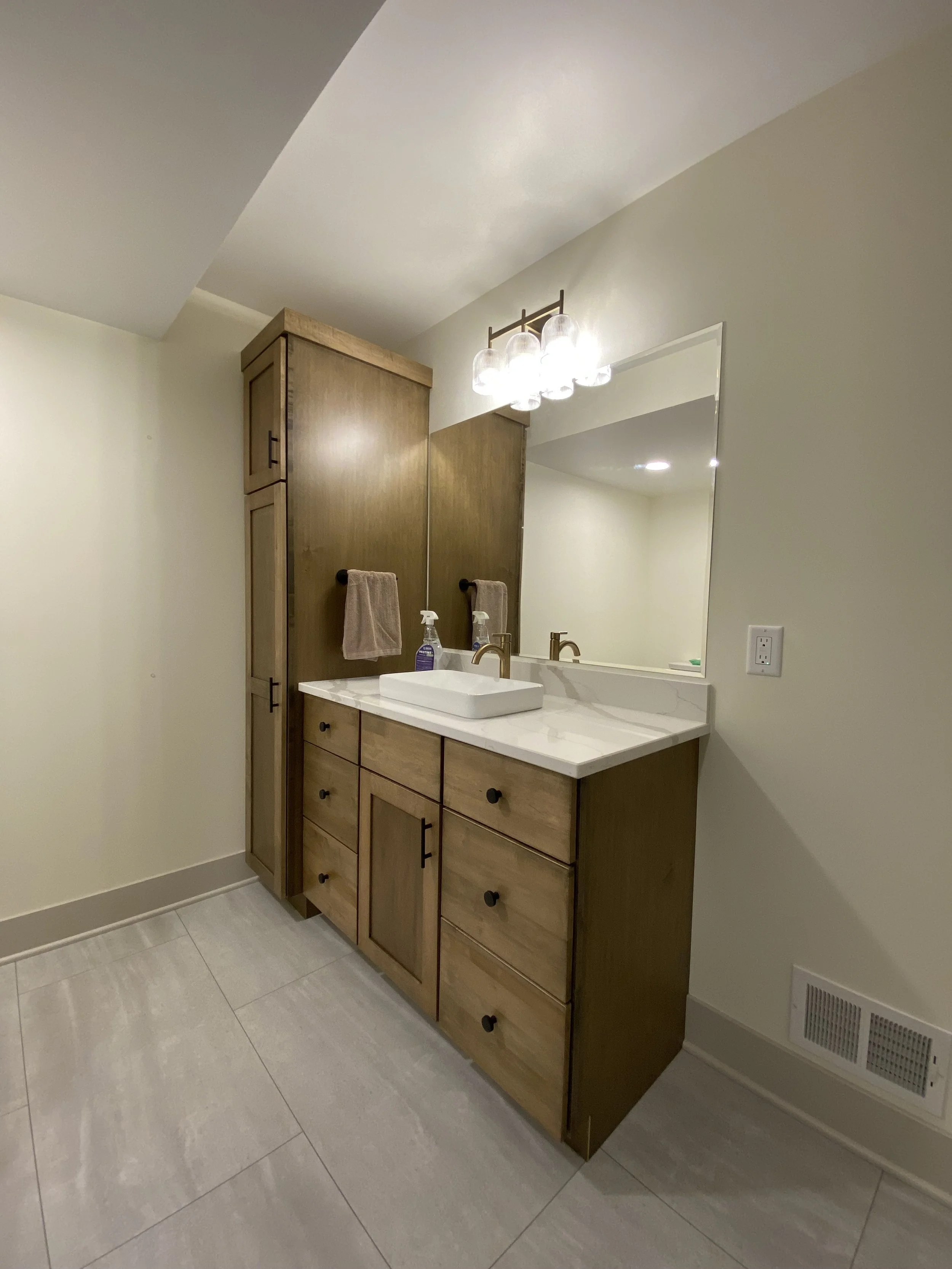 Bathroom with a wooden vanity, sink, and mirror. Overhead light fixture with bulbs. Beige wall and large floor tiles. Hand soap and towel visible.