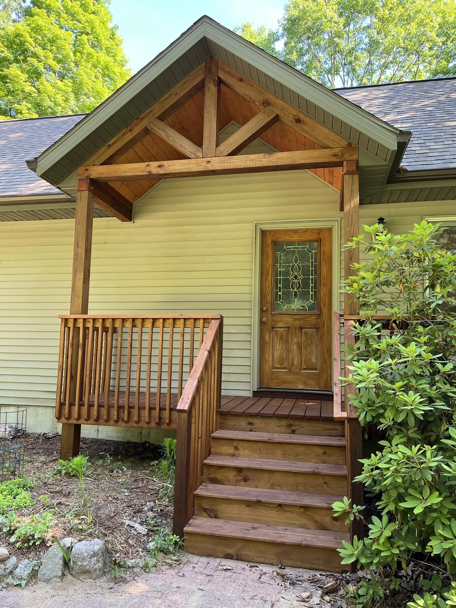 Wooden porch with stairs and railing in front of a house with beige siding, decorative glass door, and overhanging roof in a garden setting.