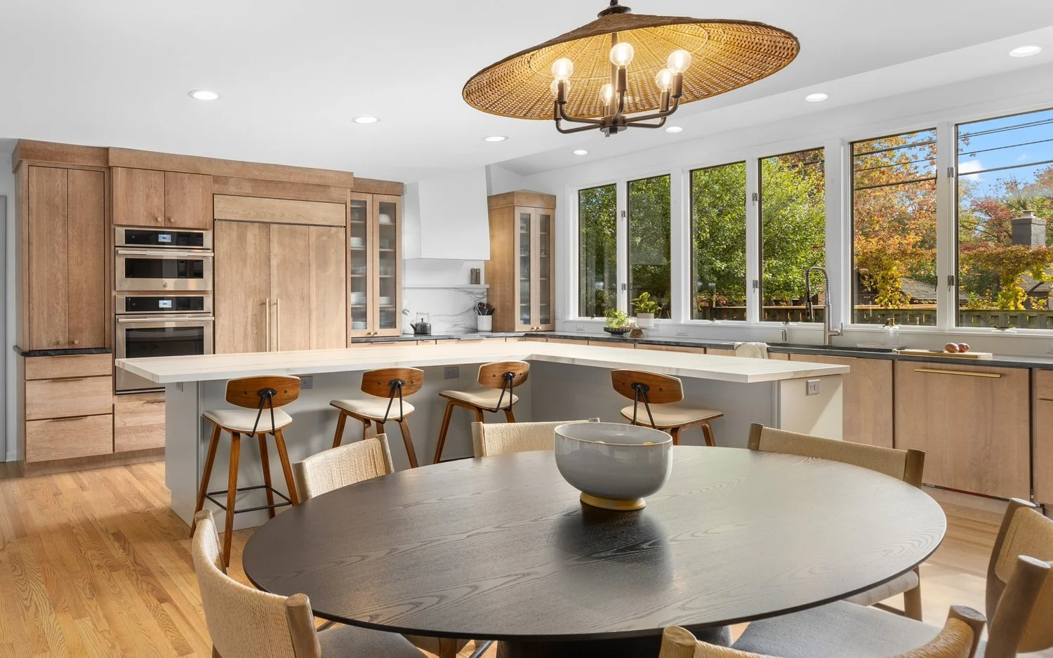 Modern kitchen with wooden cabinets, island with stools, round dining table, large windows, and decorative light fixture.
