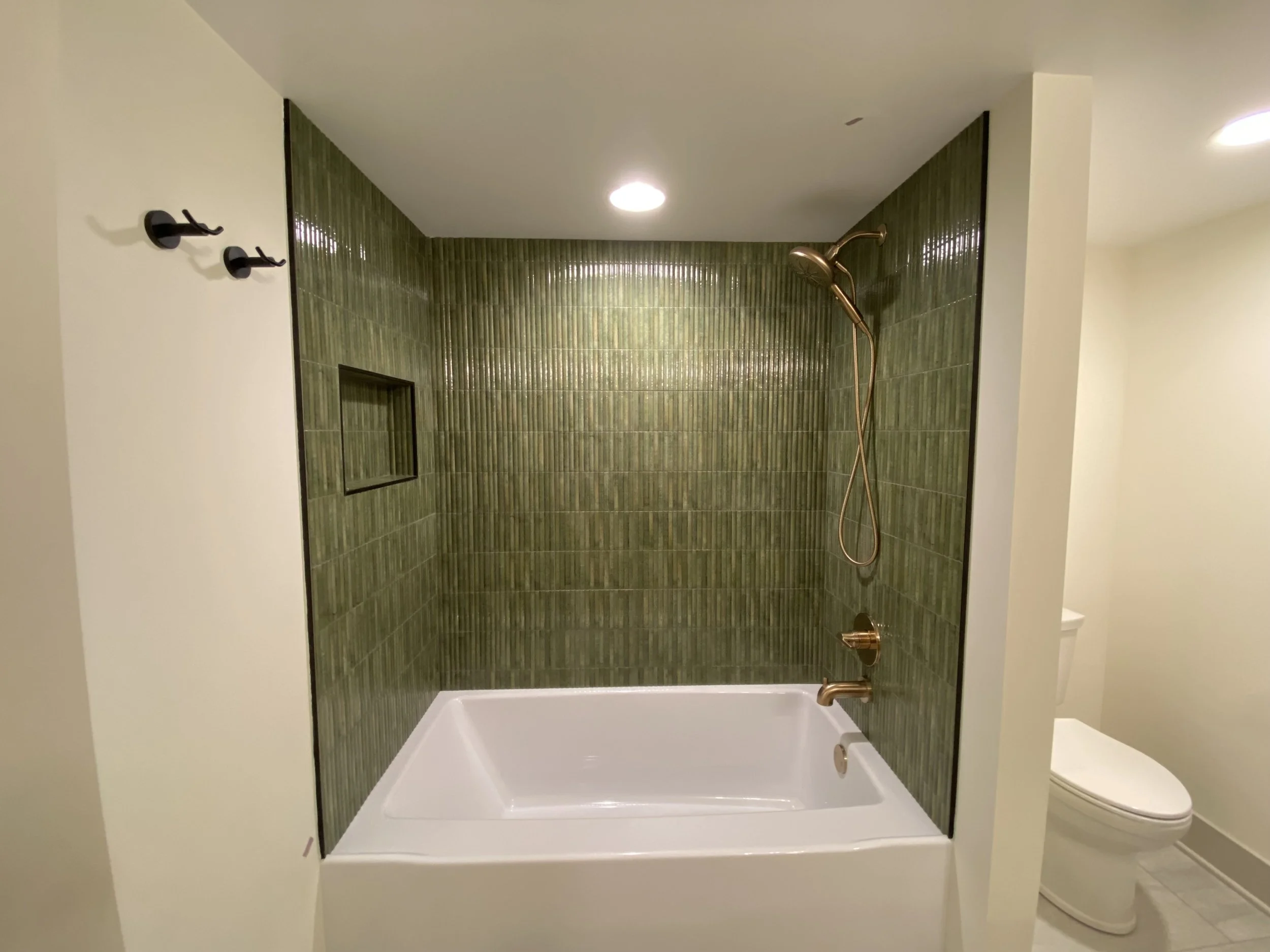 Modern bathroom with green tiled shower, white bathtub, bronze fixtures, built-in shower niche, and adjacent toilet.