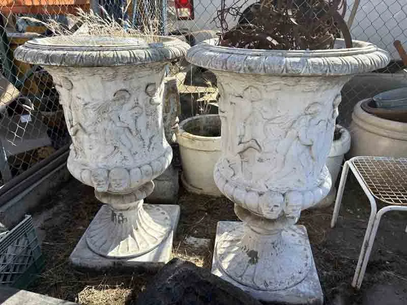 Two large, decorative, white stone planters with intricate carvings, placed outdoors among other garden pots and equipment.