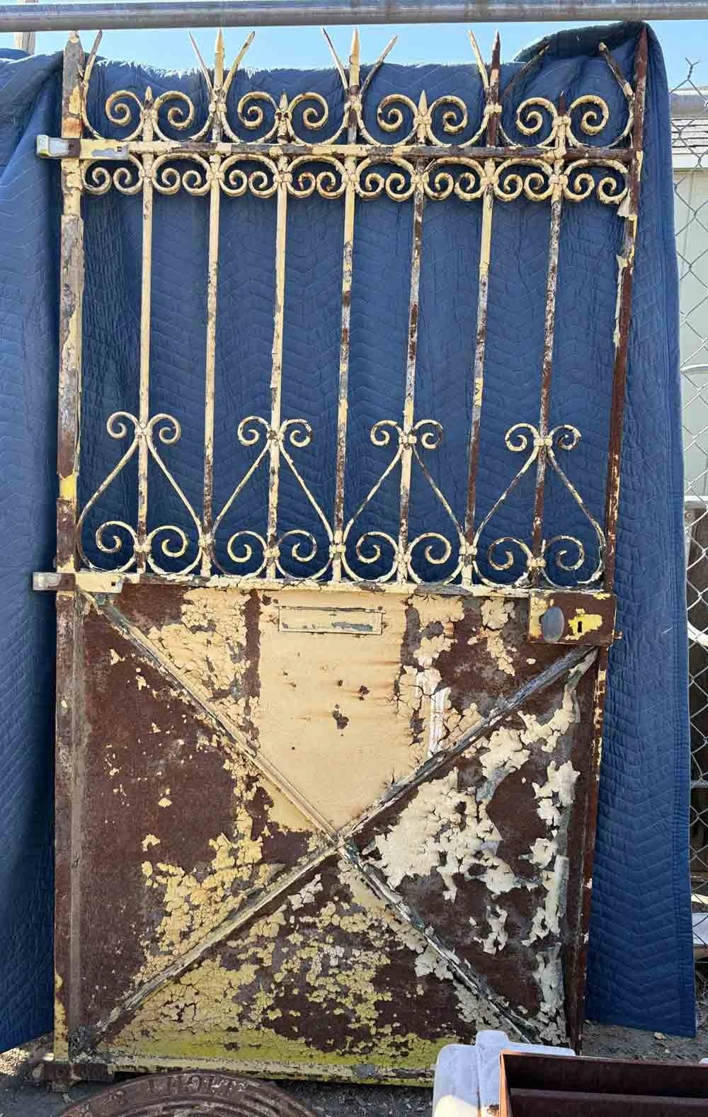 An old, rusted wrought iron gate with peeling paint, showing signs of corrosion, mounted on a blue quilted background with a chain-link fence partially visible on the right.
