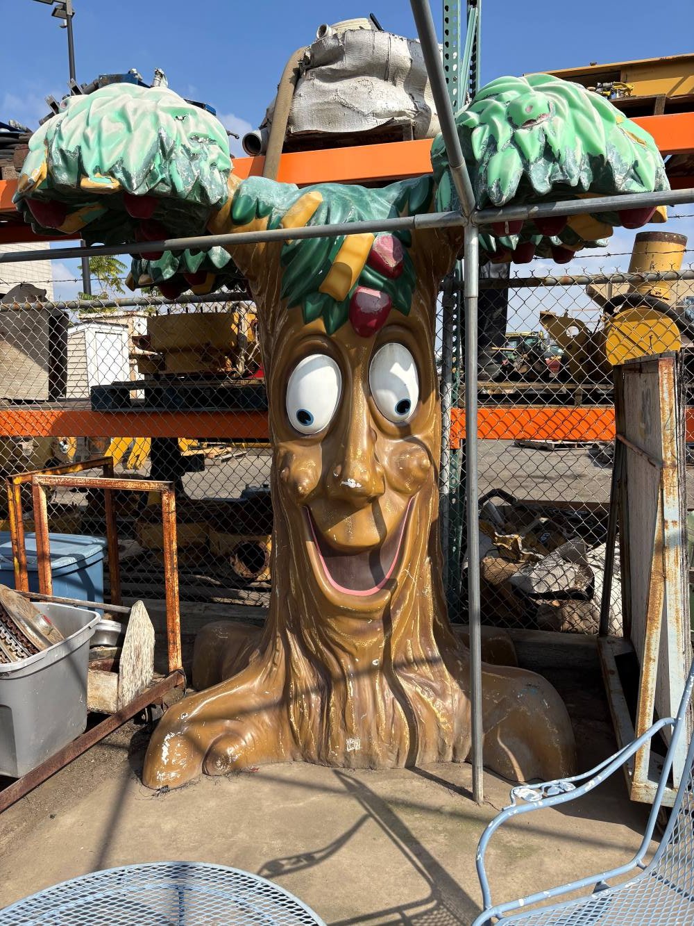 Large cartoon-style tree sculpture with a smiling face, big eyes, and green leafy canopy on top, surrounded by metal structures and outdoor items.