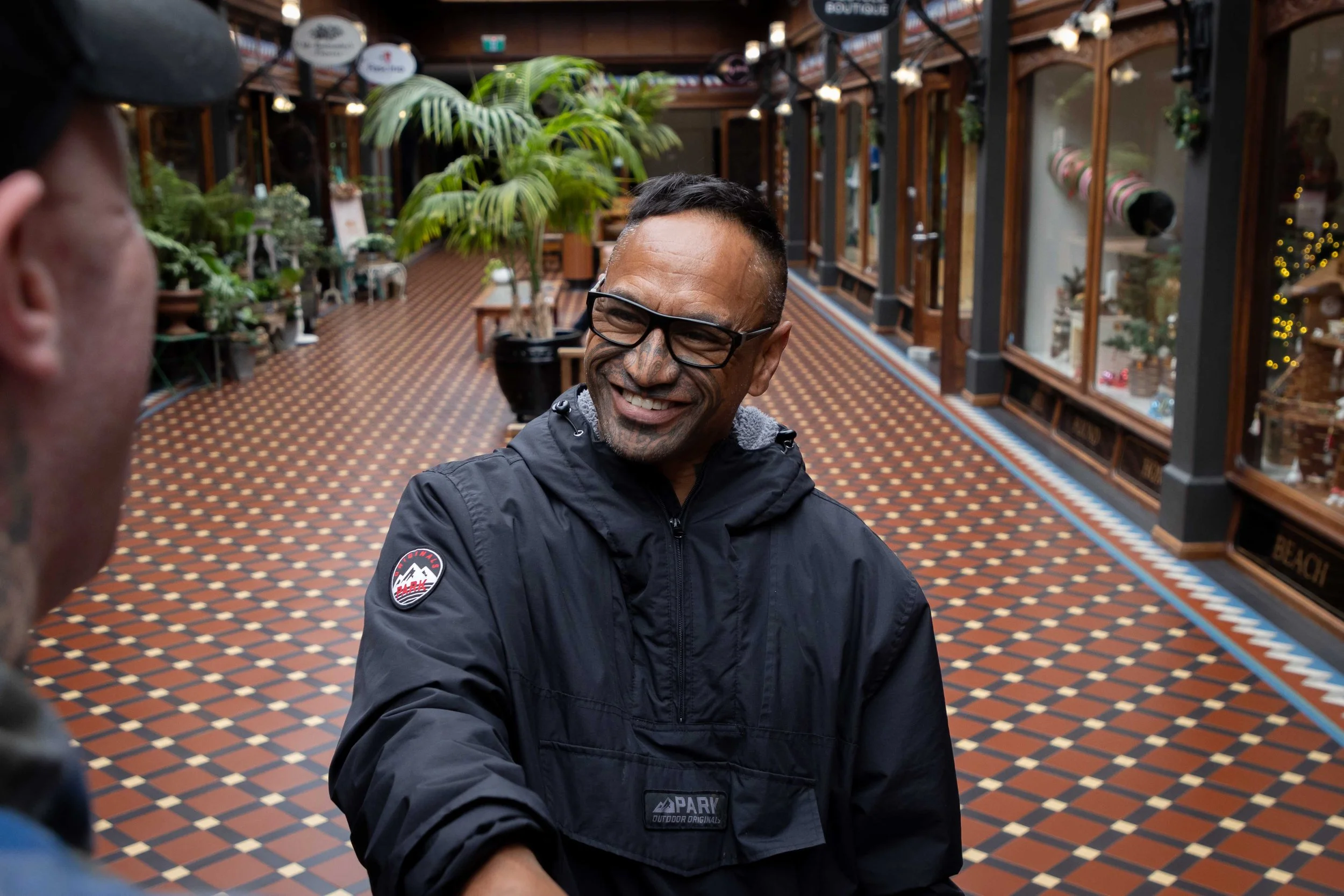 A man with glasses, short dark hair, and a gray goatee smiles and shakes hands with another person in an indoor shopping arcade filled with potted plants and display windows.