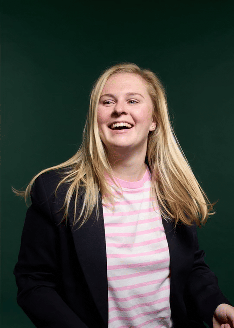 A young woman with blonde hair, smiling and looking to her left, wearing a pink and white striped shirt and a black blazer, standing against a dark green background.