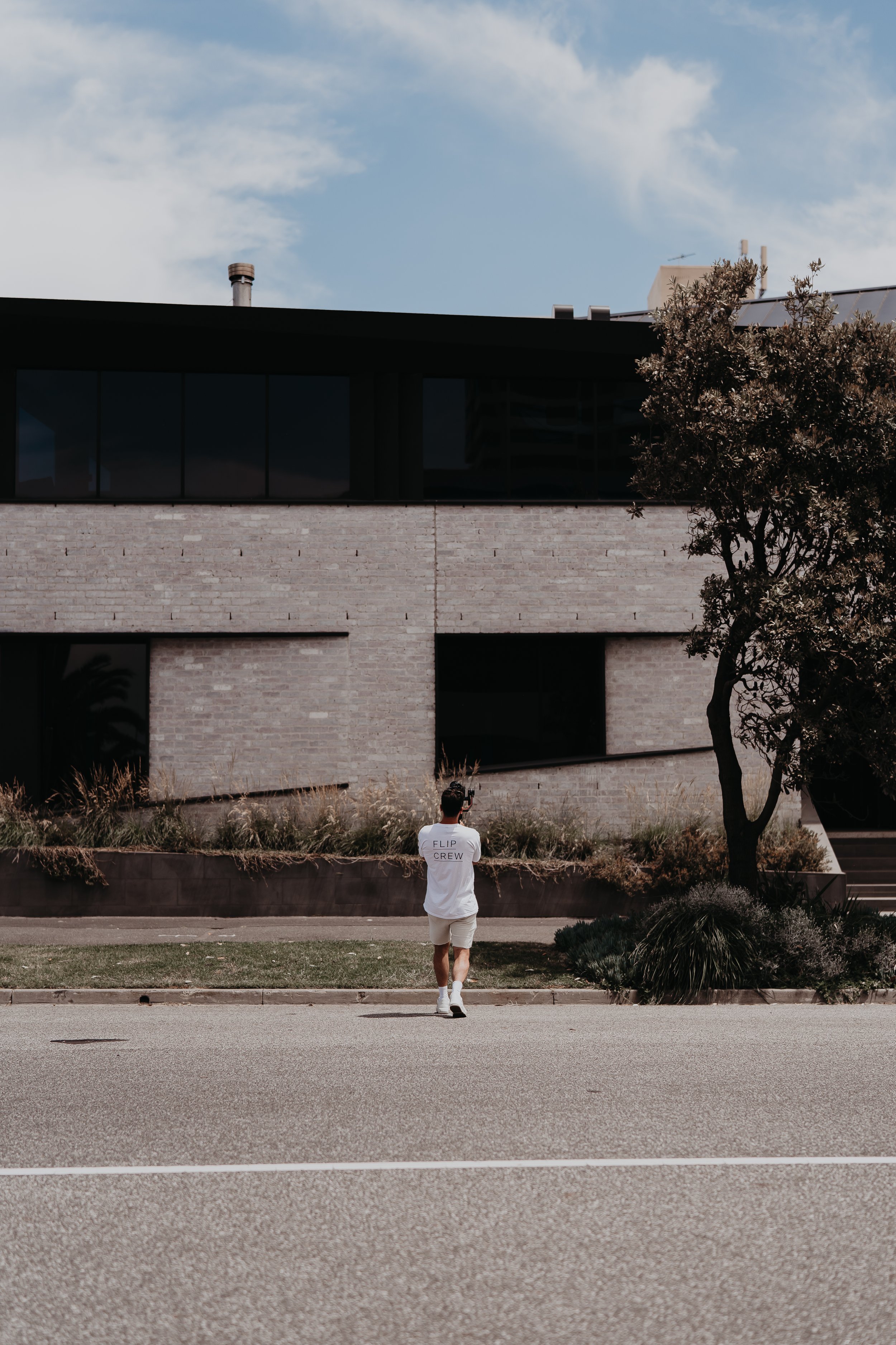 A person wearing a white T-shirt with 'Flip Crew' written on the back, shorts, and sneakers, walking on an empty road towards a modern building with large windows and a brick facade, with trees and landscaping in the foreground, under a partly cloudy sky.