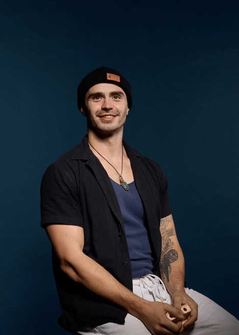 Smiling young man with tattoos, wearing a black beanie, black shirt open over a blue shirt, and a necklace, sitting against a dark blue background.