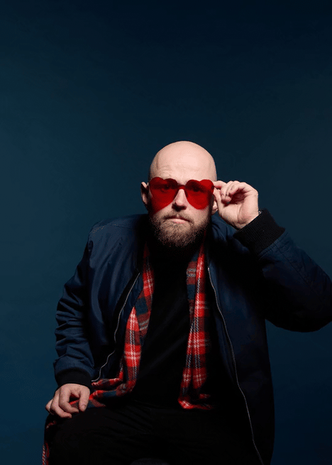 A man with a bald head and beard wearing large red sunglasses, a black jacket, and a red plaid shirt, adjusting his sunglasses against a dark blue background.