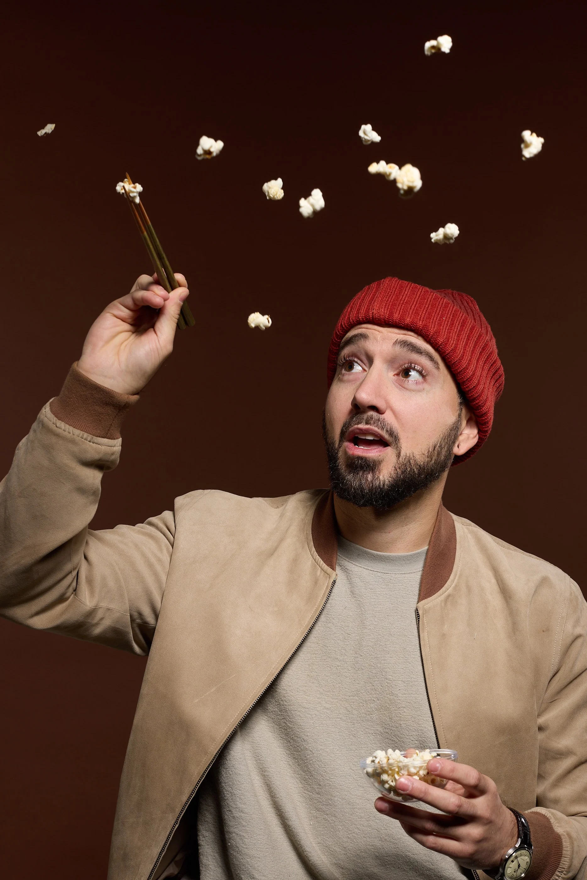 A man wearing a red knit beanie, beige jacket, and gray shirt is holding popcorn with his left hand and a brown object, possibly a spoon, in his right hand. He is looking up with an open mouth as popcorn falls around him against a brown background.