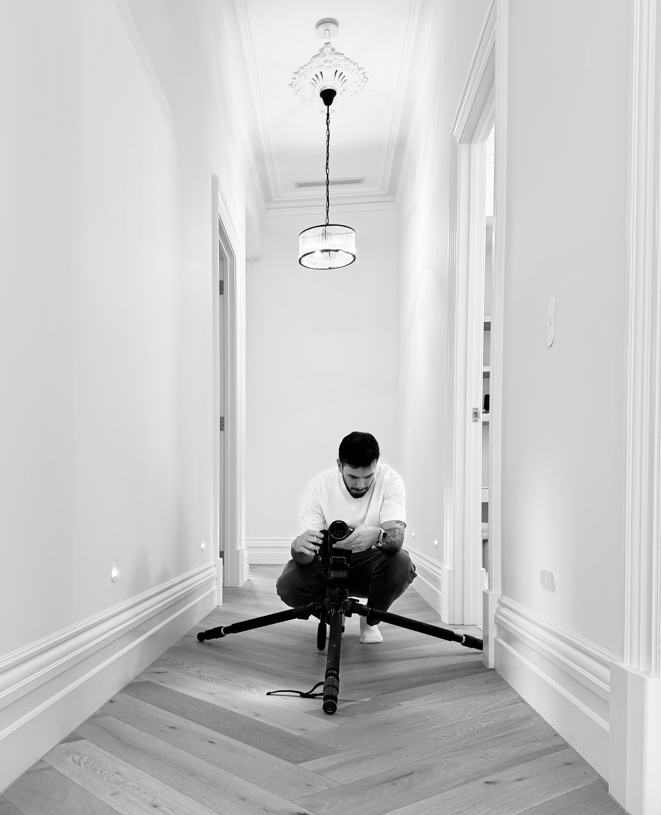 Person kneeling on a tripod-mounted camera in a hallway with white walls and decorative crown molding, with a chandelier hanging from the ceiling and a window on the right side.