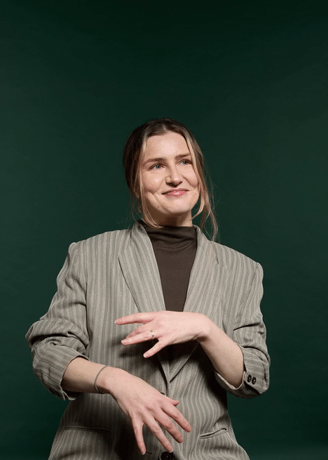 A woman with brown hair in a ponytail, wearing a gray pinstripe blazer over a brown turtleneck, smiling softly against a dark green background.