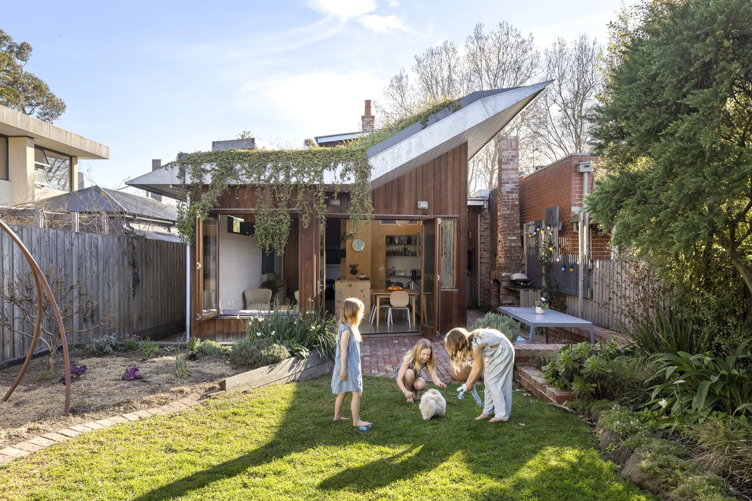 Children playing on a garden lawn, with a modern wooden house in the background, featuring large open doors, a lounge area, and a dining table. The scene is outdoors with trees and plants, under a clear blue sky.