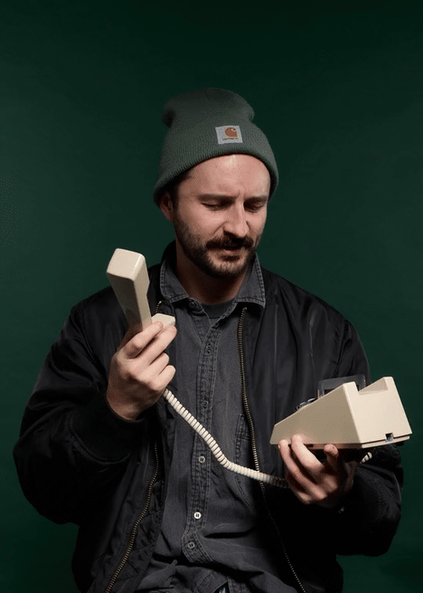 A man wearing a gray beanie and black jacket holding a vintage beige telephone receiver and base, looking confused or concerned, against a green background.