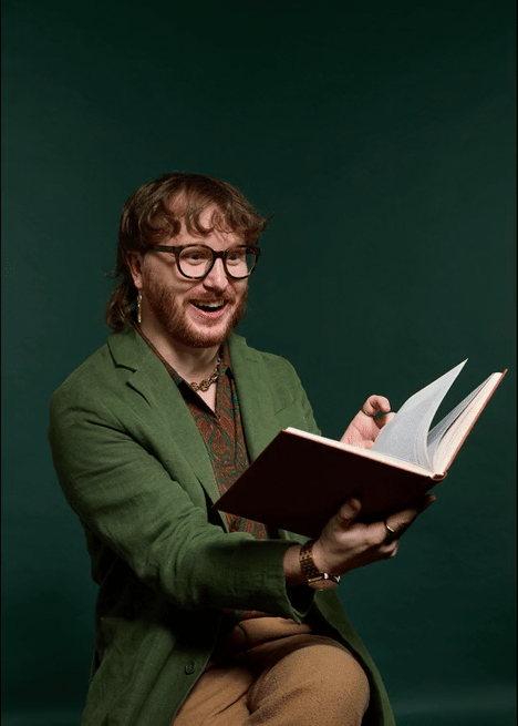 A man with glasses, a beard, and long hair, wearing a green blazer and patterned shirt, sitting and reading a book, smiling against a dark green background.