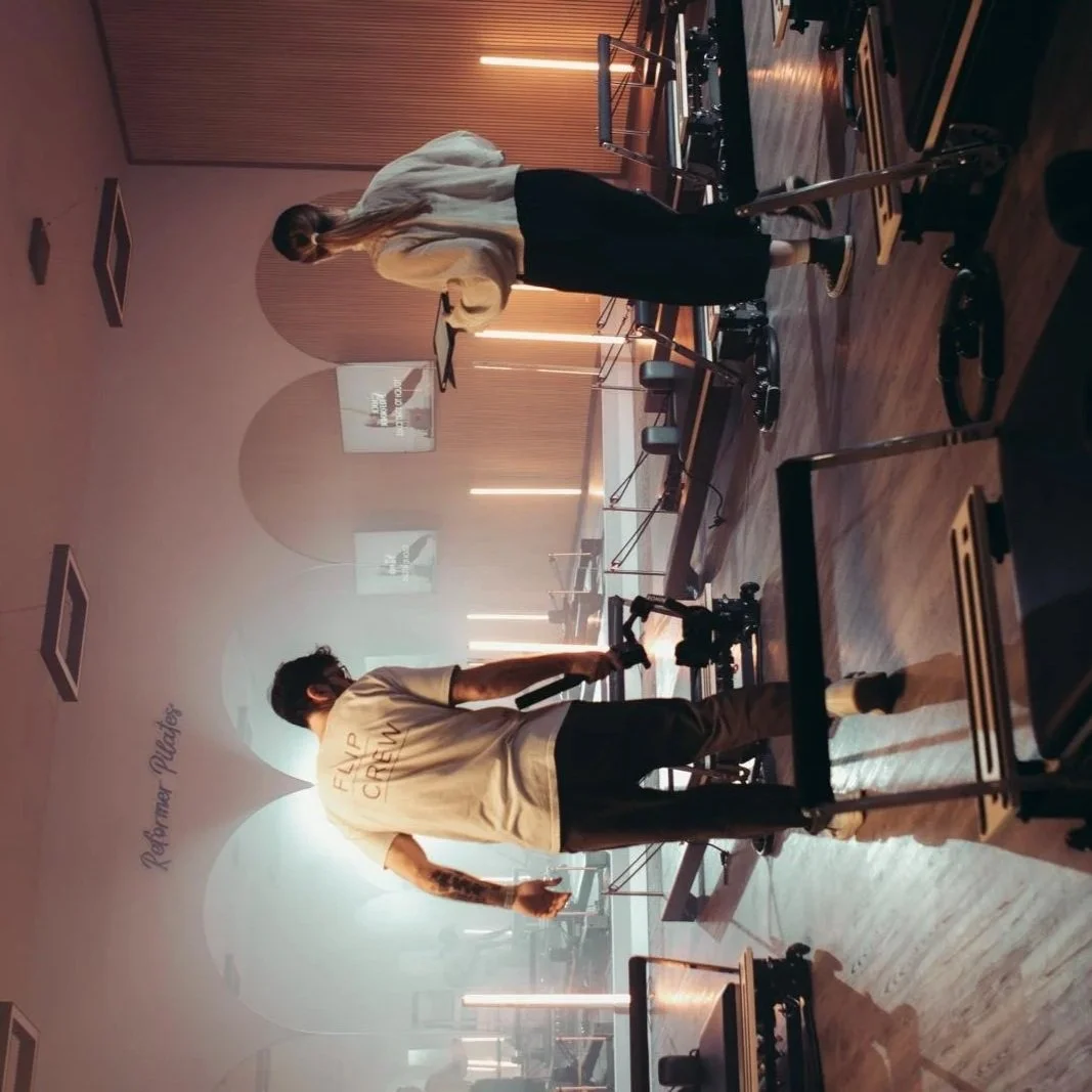 A woman standing in a pilates studio, holding a tablet, and a man filming using a camera on a gimbal. There are exercise equipment and a projector screen in the background. Filming video content for YourReformer