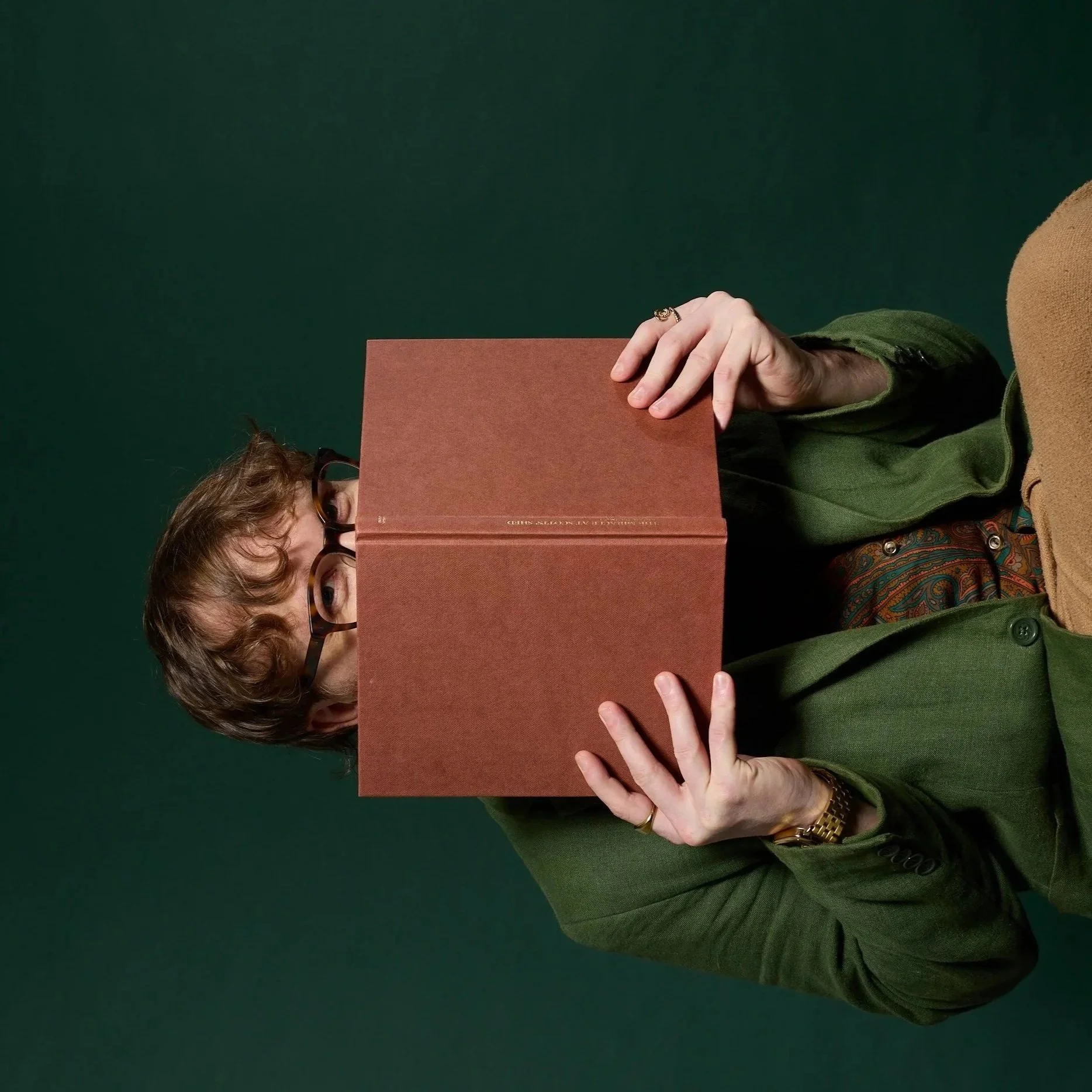 A person with curly hair, glasses, and a watch holding a large brown book in front of their face, wearing a layered outfit with green, brown, and patterned fabrics, against a dark green background.