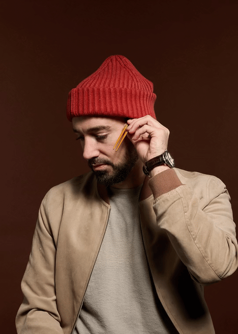 Man wearing a red knit beanie, beige jacket, light gray shirt, holding a yellow pencil to his temple, looking downward with a thoughtful expression, dark background.