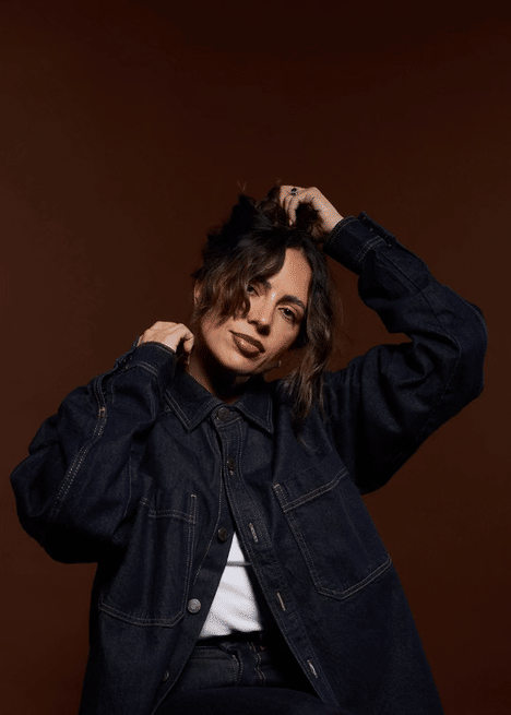 Woman with wavy hair in a denim jacket posing against a brown background.