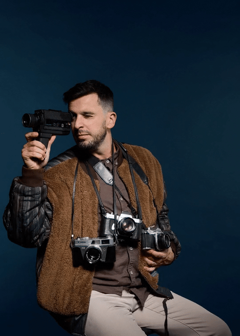 Man with multiple cameras hanging around neck holding a camera to his eye against a dark background.
