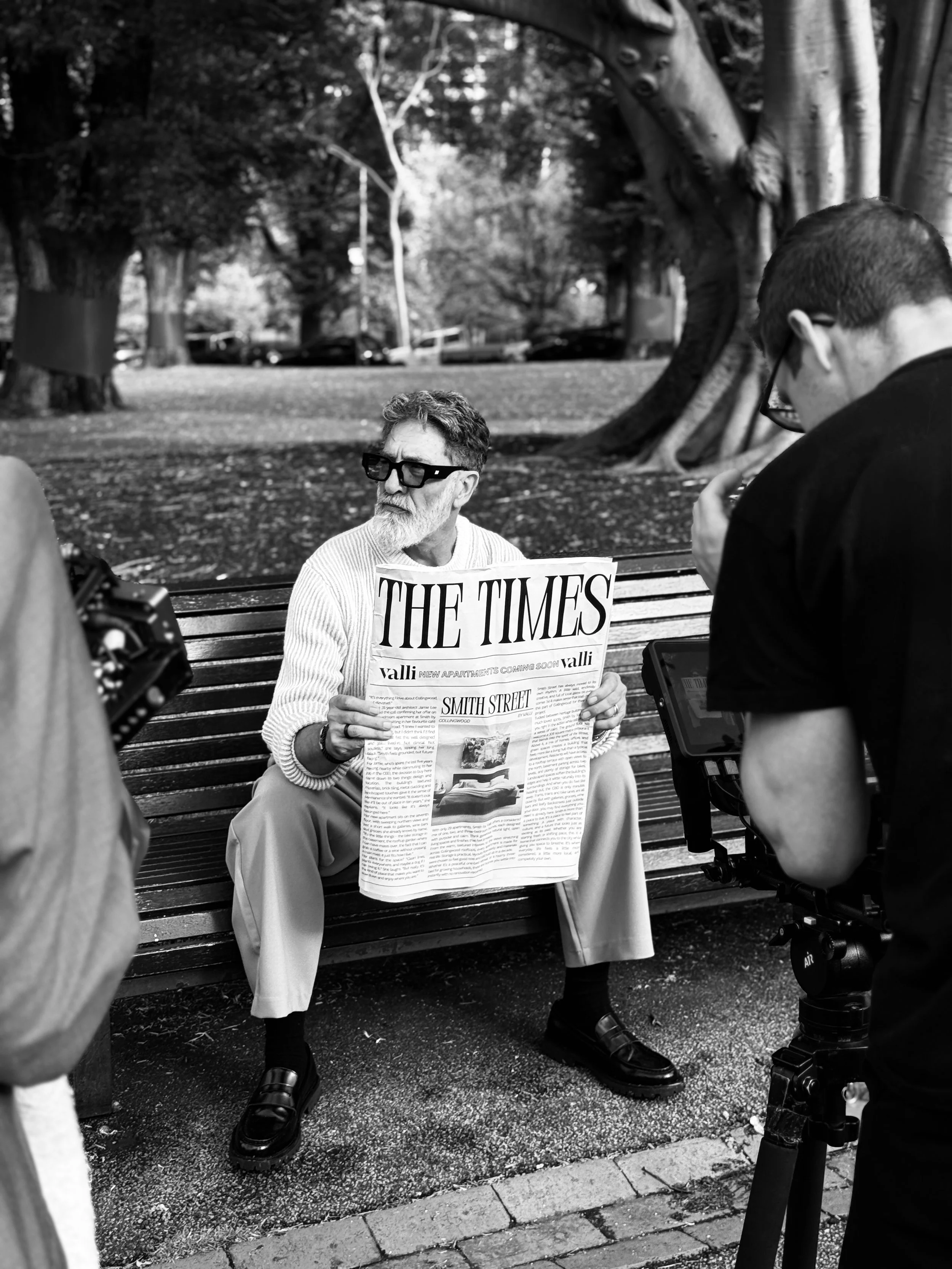 A man with glasses and a beard, seated on a park bench, holding a newspaper titled 'The Times'. He is surrounded by two other people, one partially visible on the left and another standing on the right, who appear to be filming or photographing him. The scene is outdoors with large trees and parked cars in the background.