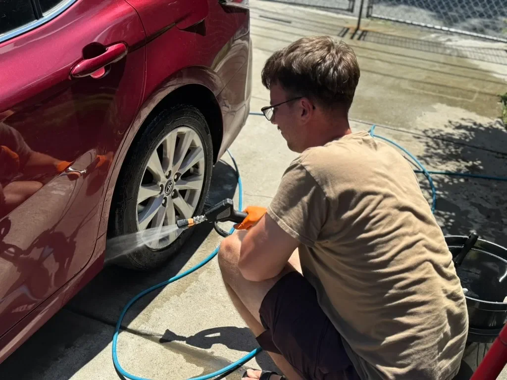 A man wearing glasses, a tan t-shirt, and shorts is washing or rinsing the wheel of a red car with a garden hose. He is squatting on the driveway next to a bucket and a blue hose.