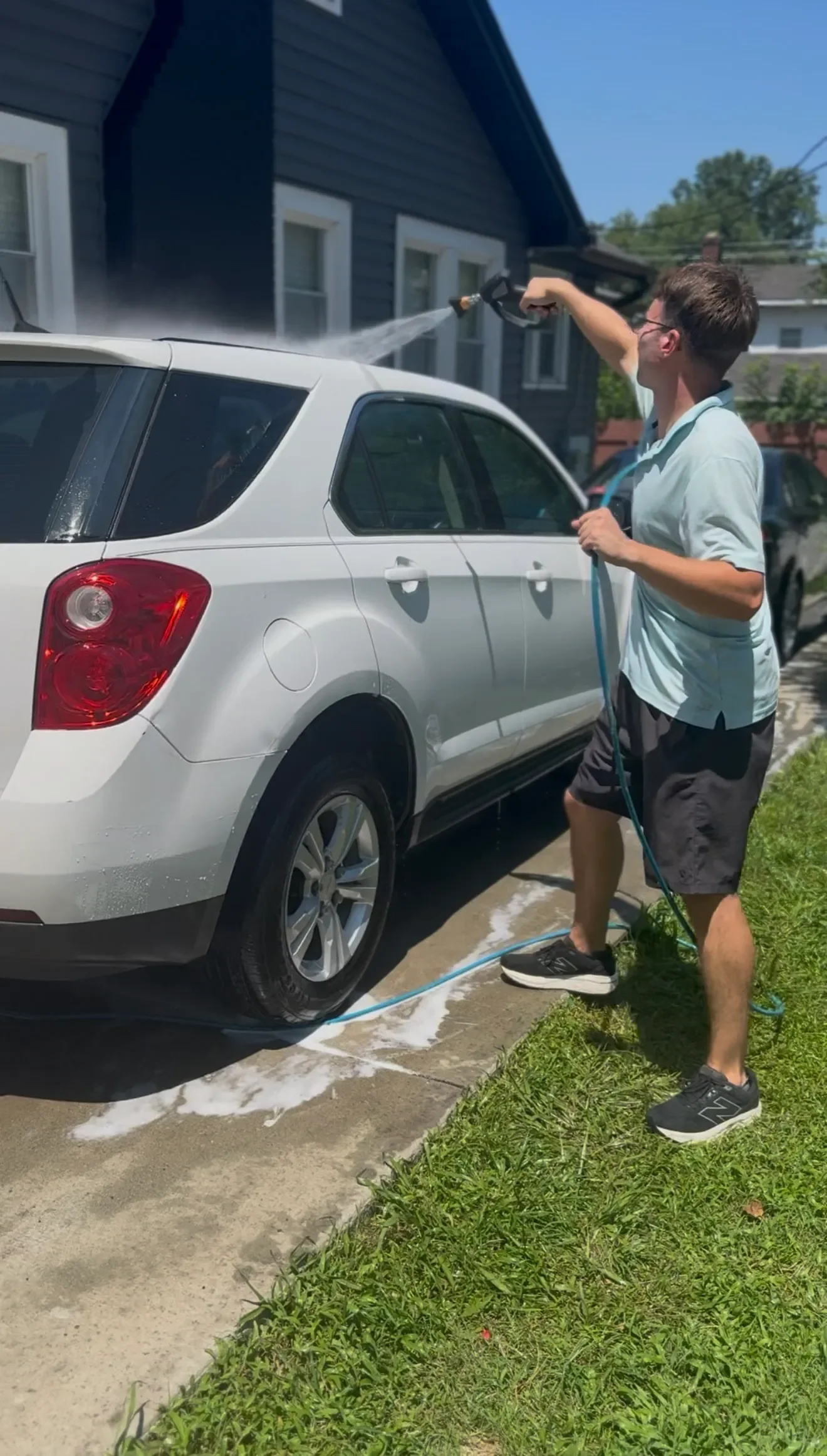 A man wearing glasses, a light blue shirt, and black shorts is washing a white SUV with a hose, standing on the driveway next to a grassy lawn on a sunny day.