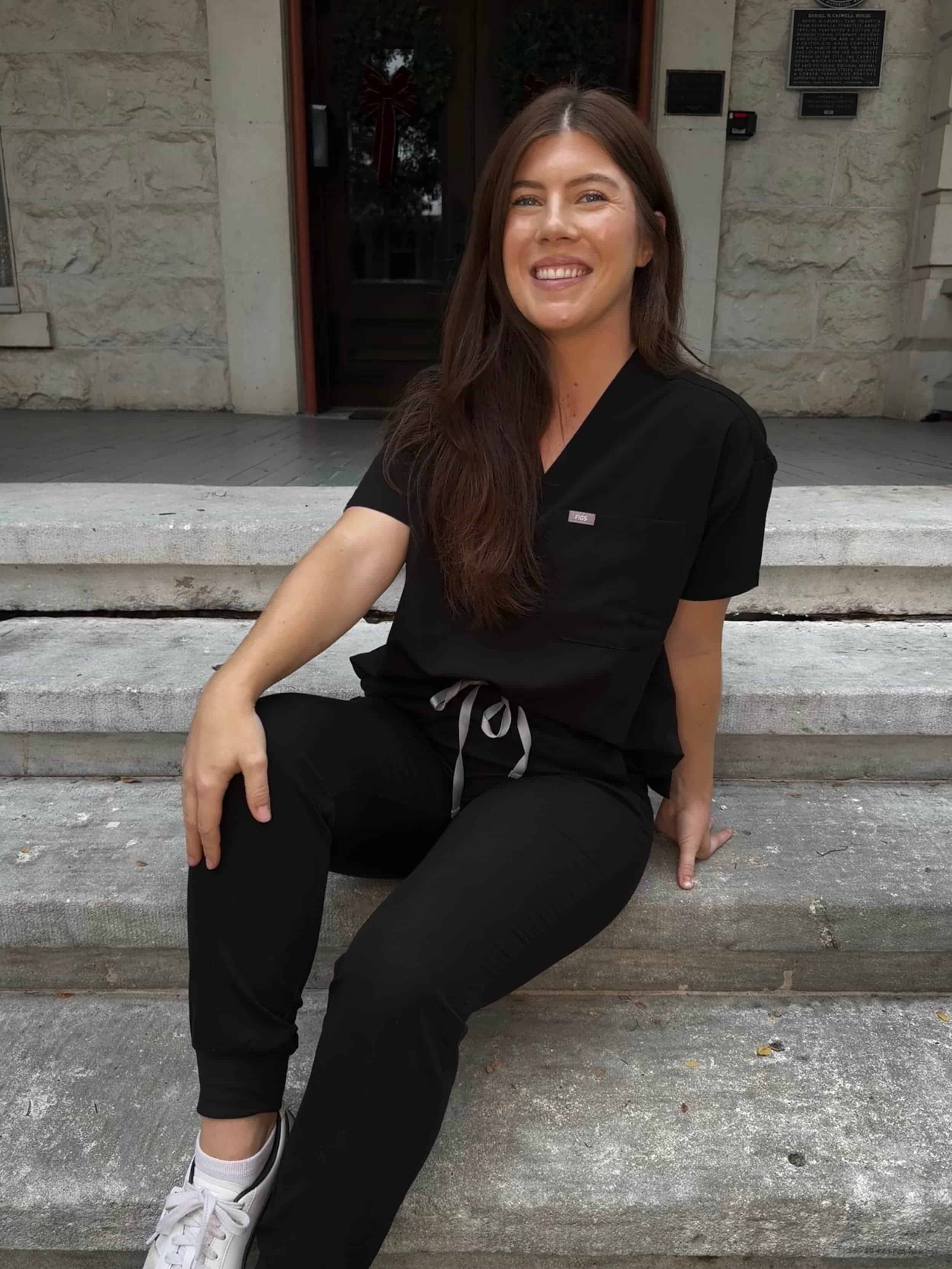 Woman with long dark hair sitting on stone steps outside a building, smiling, wearing black scrubs and white sneakers.